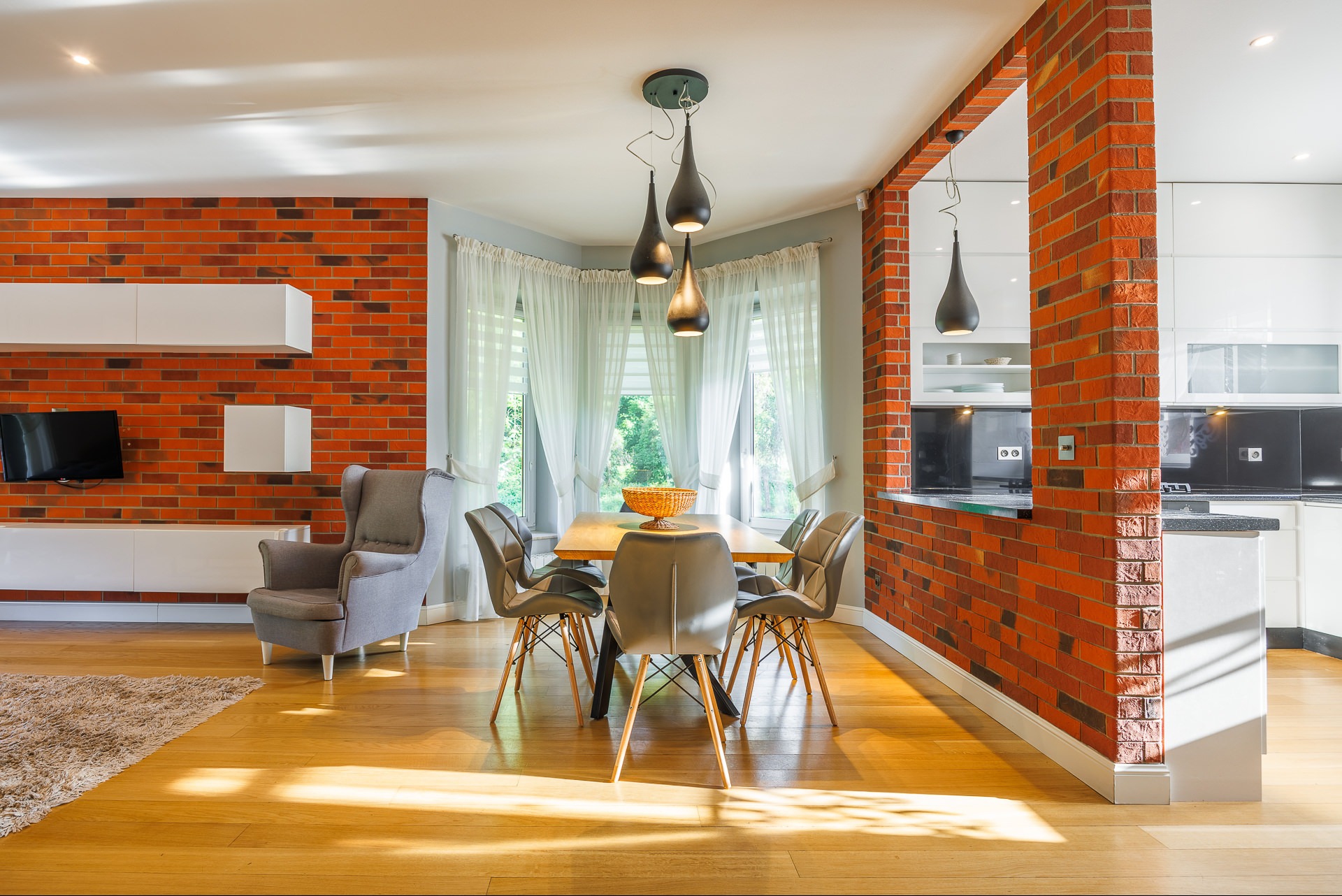 Jasne wnętrze z ceglaną ścianą, drewnianym stołem i krzesłami. Widoczny fragment kuchni z czarnym blatem. Nowoczesny design, ciepłe oświetlenie.