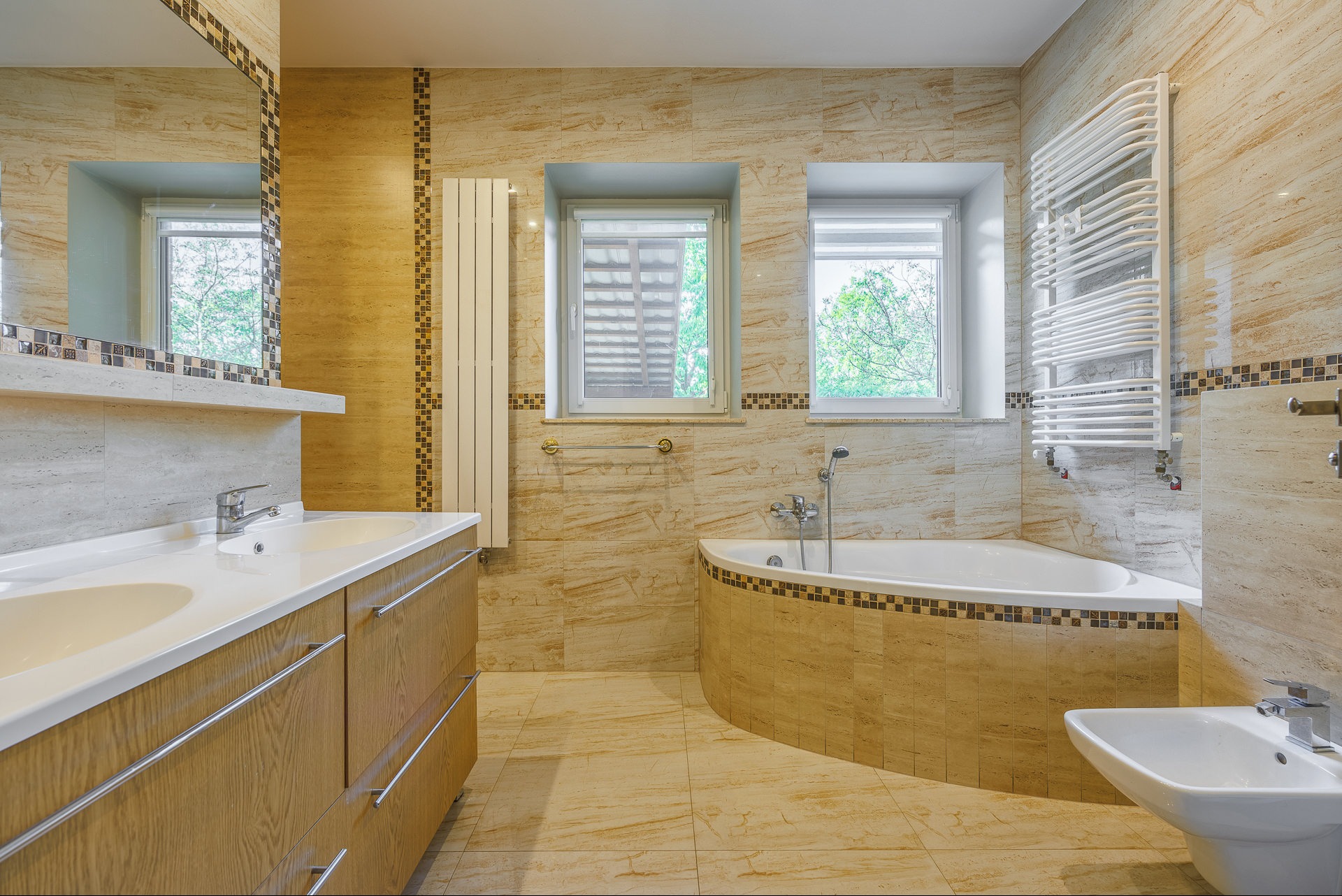 Jasna łazienka z beżowymi płytkami imitującymi marmur, z wanną narożną, umywalką z szafką i bidetem. Dwa okna z widokiem na zieleń. Grzejnik drabinkowy.