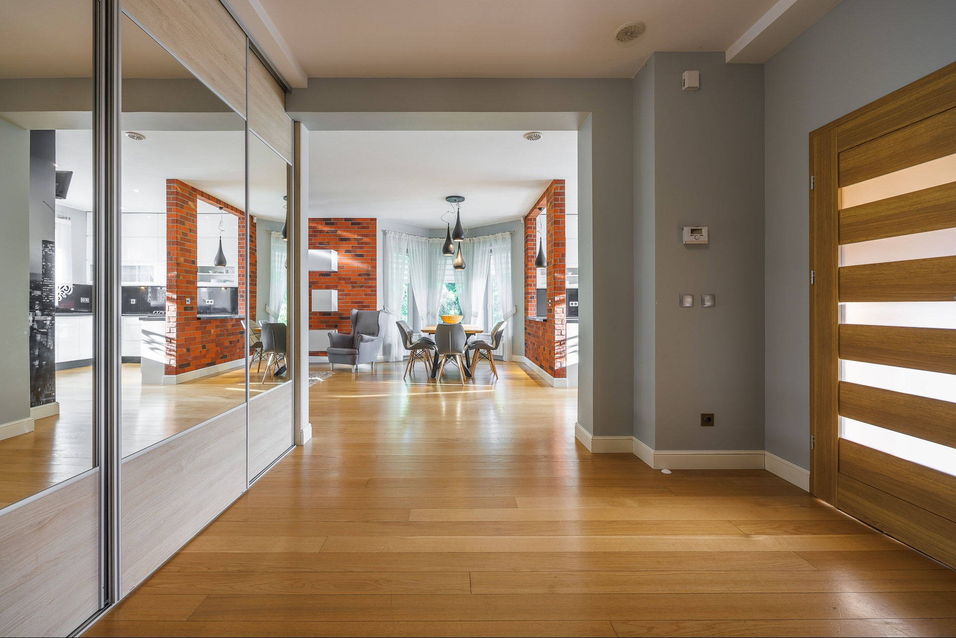 Nowoczesne wnętrze z parkietem, ceglaną ścianą i lustrzaną szafą. Widok na jadalnię z szarymi krzesłami i lampami wiszącymi. Jasne, przestronne pomieszczenie.