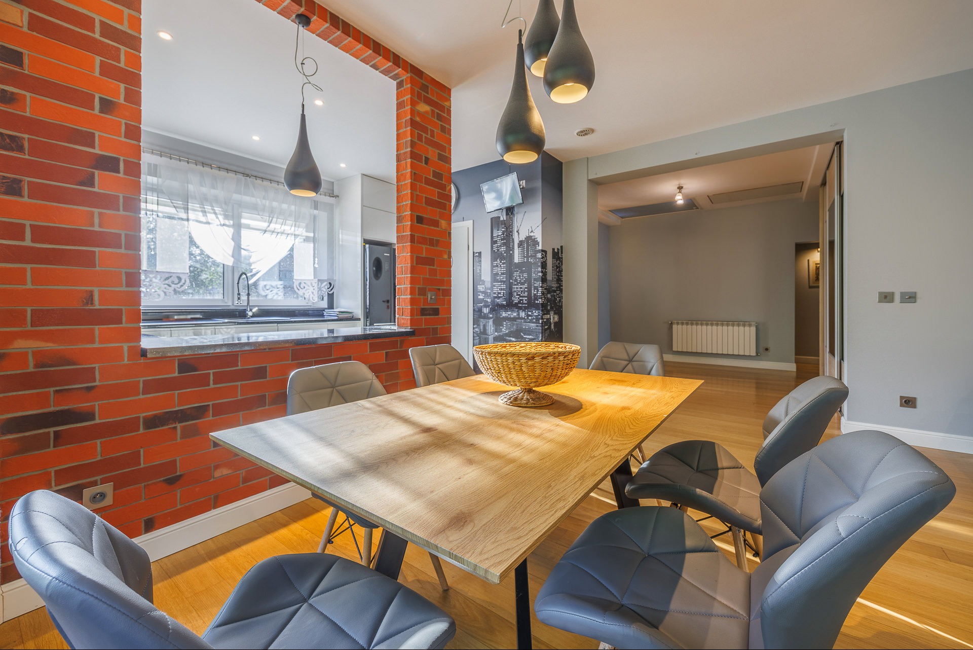 Nowoczesna jadalnia z drewnianym stołem, szarymi krzesłami i ceglaną ścianą z oknem na kuchnię. Nad stołem wiszą trzy designerskie lampy.