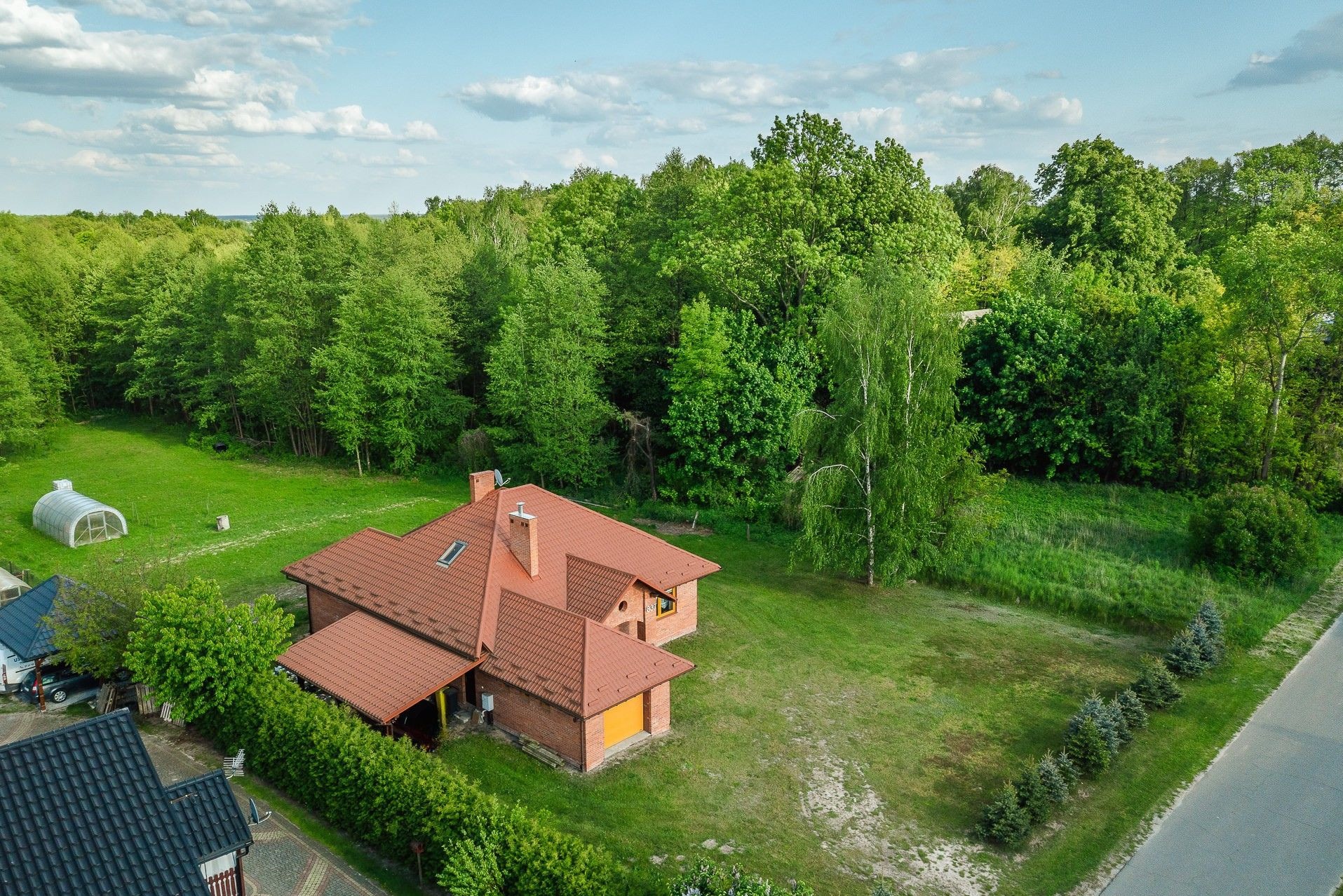 Zdjęcie z drona przedstawia dom z czerwoną dachówką otoczony zielenią, z widocznym ogrodem, lasem w tle i fragmentem drogi po prawej stronie kadru.