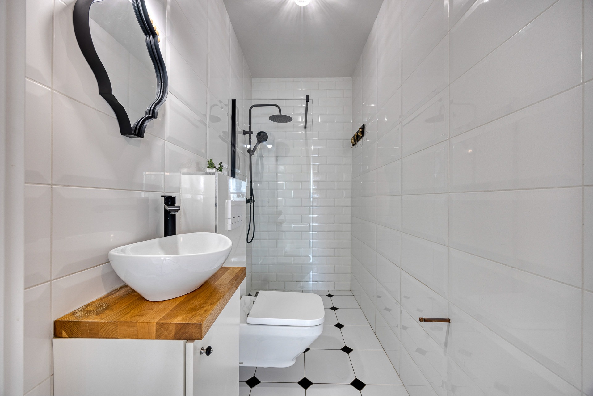 Biała łazienka z umywalką na drewnianym blacie, czarną armaturą i kabiną prysznicową. Minimalistyczny design z akcentami czerni i drewna.