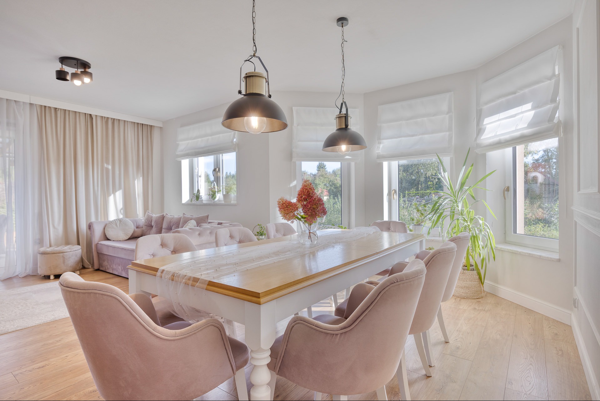Jasne wnętrze jadalni z drewnianym stołem, różowymi krzesłami i sofą w tle. Dwa wiszące, metalowe klosze oświetlają stół. Okna z roletami rzymskimi.