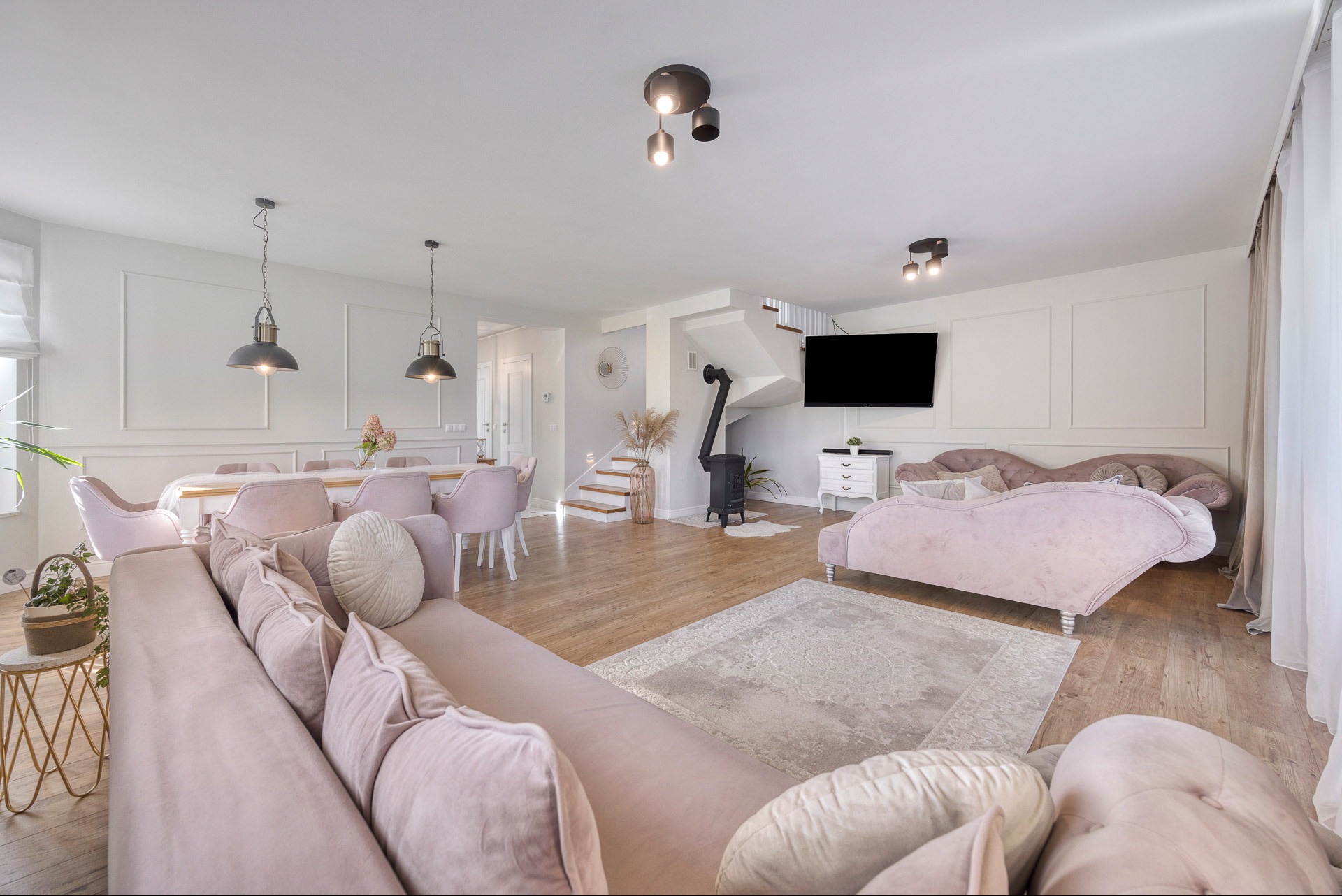 Jasne wnętrze salonu z różowymi meblami: sofa, krzesła, szezlong. W tle kominek, schody i stół. Nowoczesne oświetlenie punktowe i wiszące lampy.