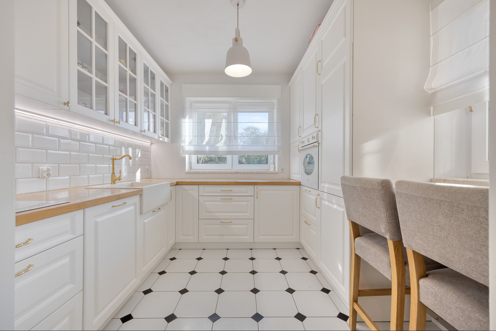 Biała kuchnia w stylu klasycznym z drewnianymi blatami, złotą armaturą i czarno-białą podłogą. Widoczne krzesła barowe i okno z roletą rzymską.