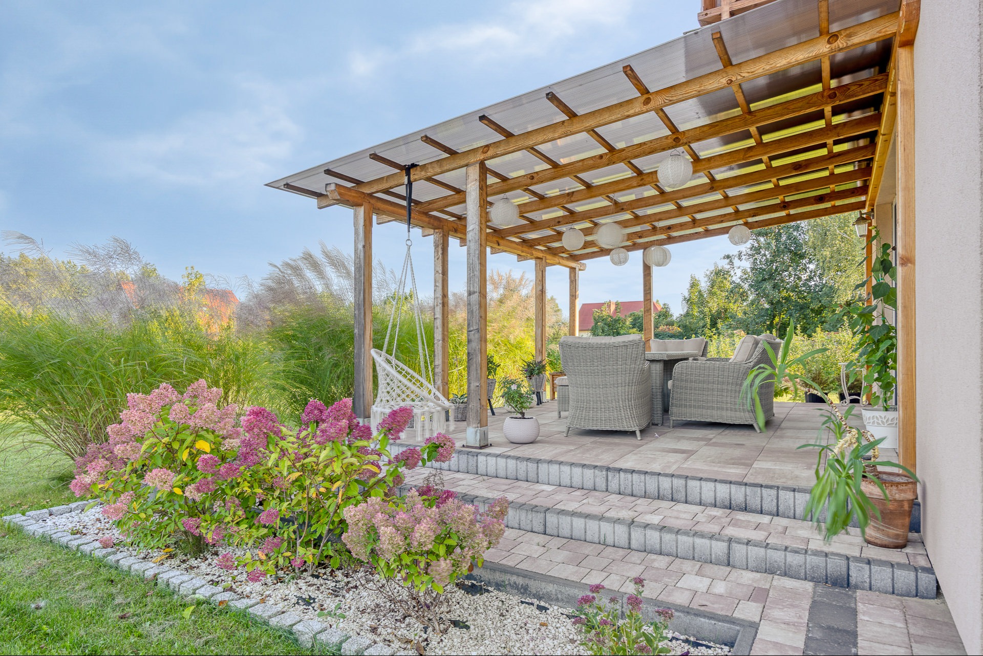 Zadaszony taras z drewnianą konstrukcją, rattanowymi meblami i wiszącym fotelem. Hortensje w pierwszym planie. Ujęcie w słoneczny dzień.