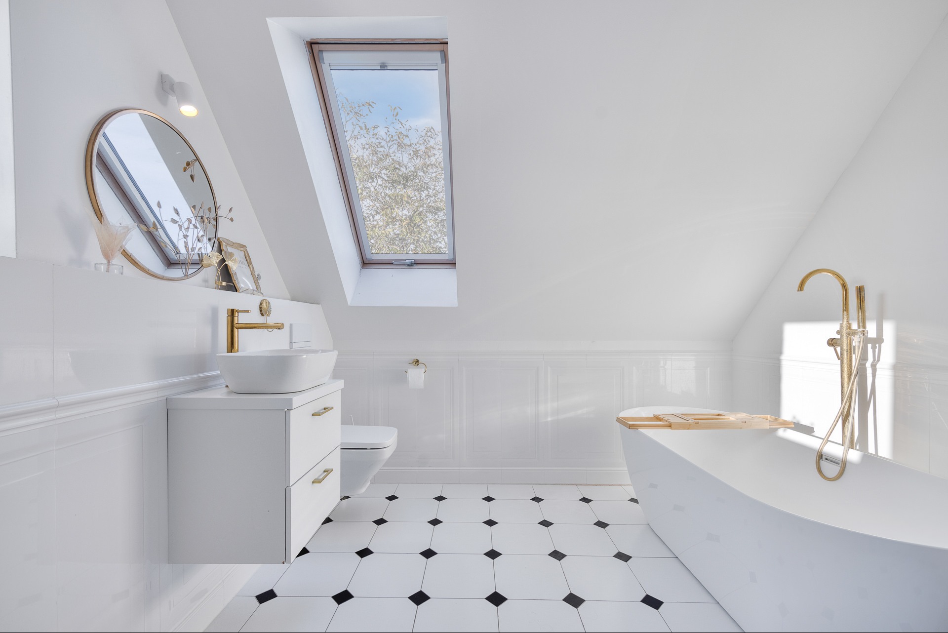 Jasna łazienka na poddaszu z oknem dachowym, białą armaturą i czarno-białą podłogą w karo. Złote akcenty w bateriach i lustrze dodają elegancji.