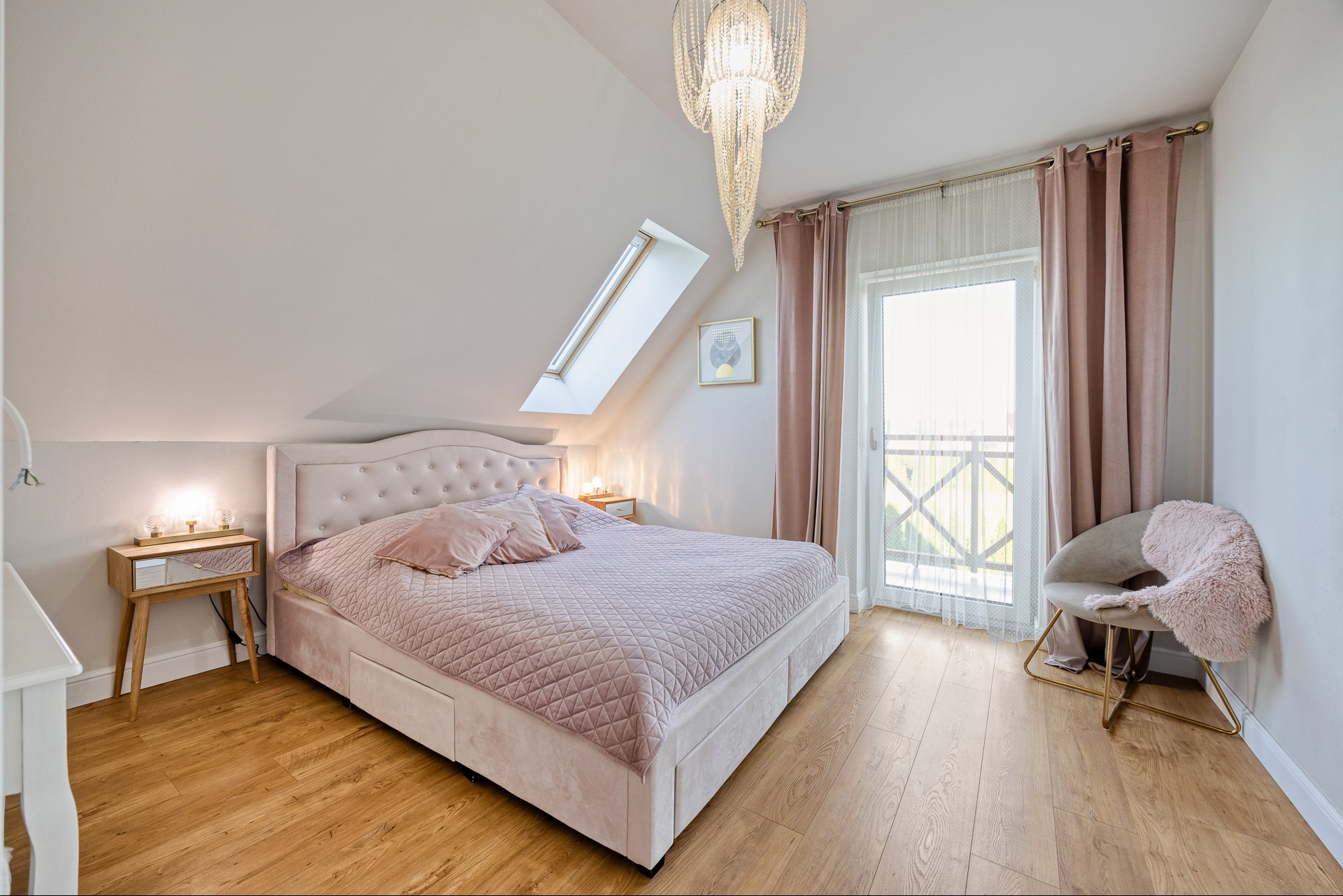 Jasna sypialnia na poddaszu z łóżkiem w pudrowym różu, drewnianą podłogą i balkonem. Kryształowy żyrandol i okno dachowe doświetlają wnętrze.