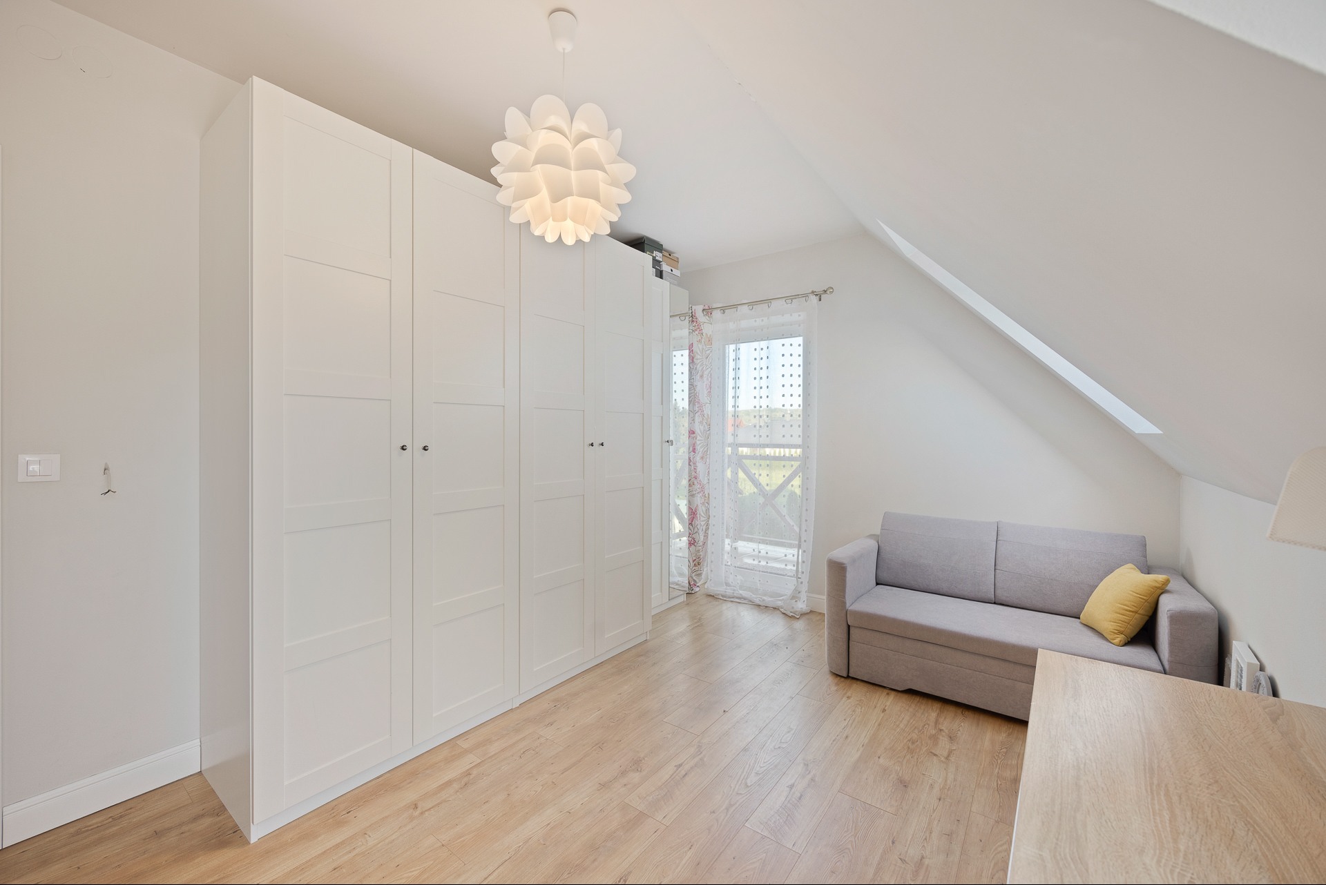 Jasne poddasze z oknem balkonowym: biała szafa w zabudowie, szara sofa z żółtą poduszką, drewniana podłoga i designerska lampa. Minimalistyczny styl.