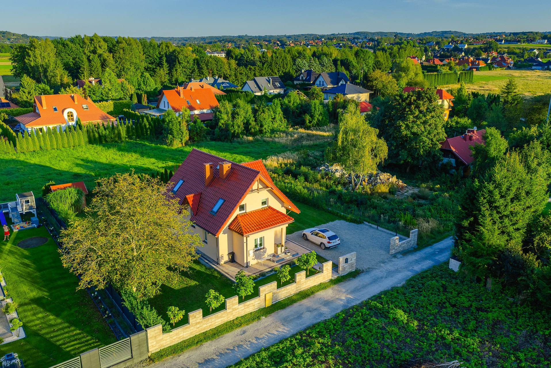 Dom jednorodzinny z czerwonym dachem, widok z lotu ptaka, otoczony zielenią i innymi domami. Na podjeździe stoi biały samochód. Słoneczny dzień.