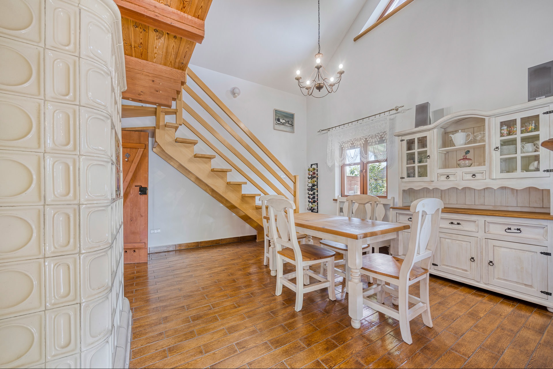 Jasne wnętrze domu z drewnianymi schodami, stołem i kredensem w stylu rustykalnym. Widoczny piec kaflowy i drewniana podłoga. Ujęcie z szerokim kątem.