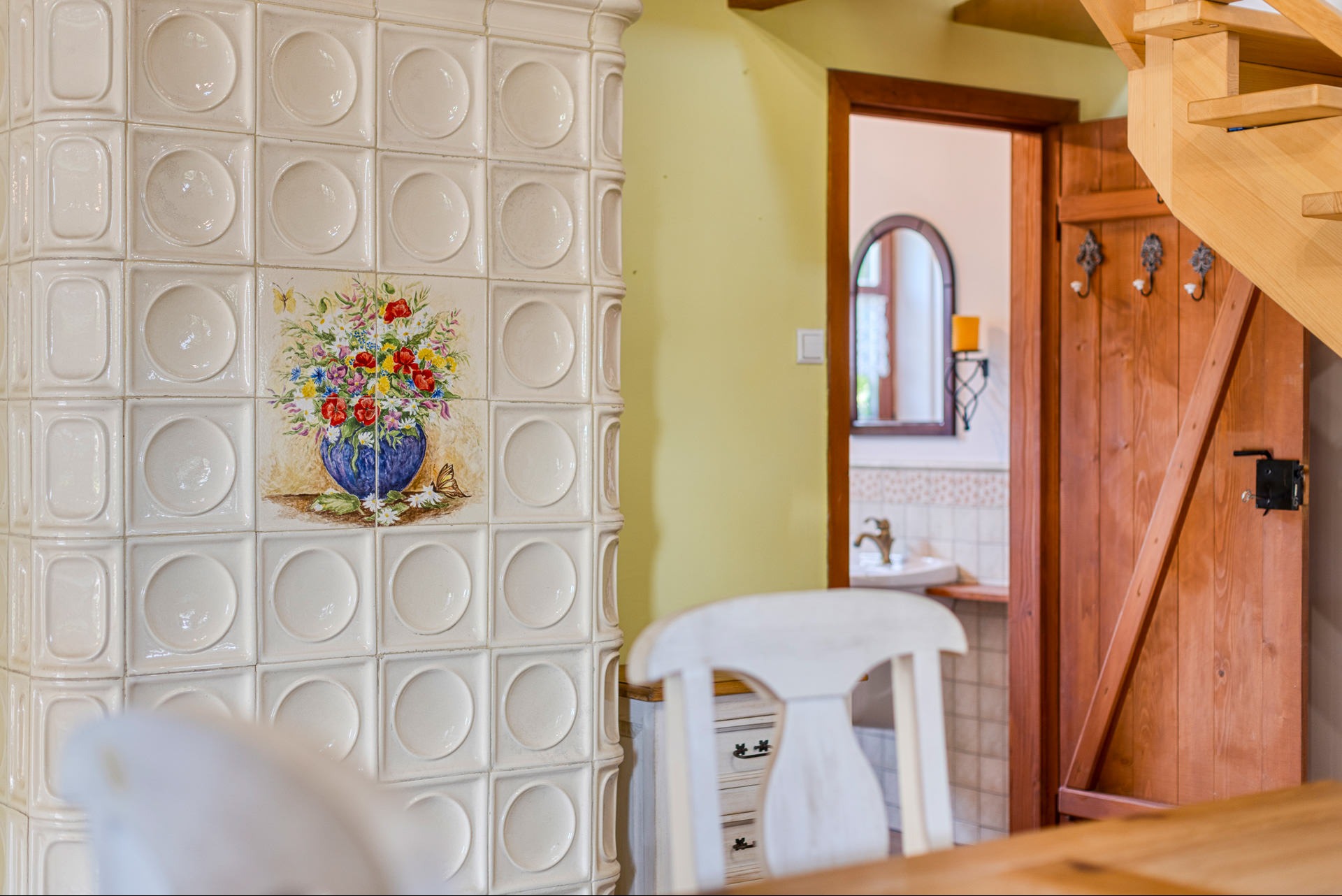 Biały piec kaflowy z motywem kwiatowym w niebieskim wazonie, widoczny fragment drewnianych schodów i drzwi do łazienki z lustrem w tle.