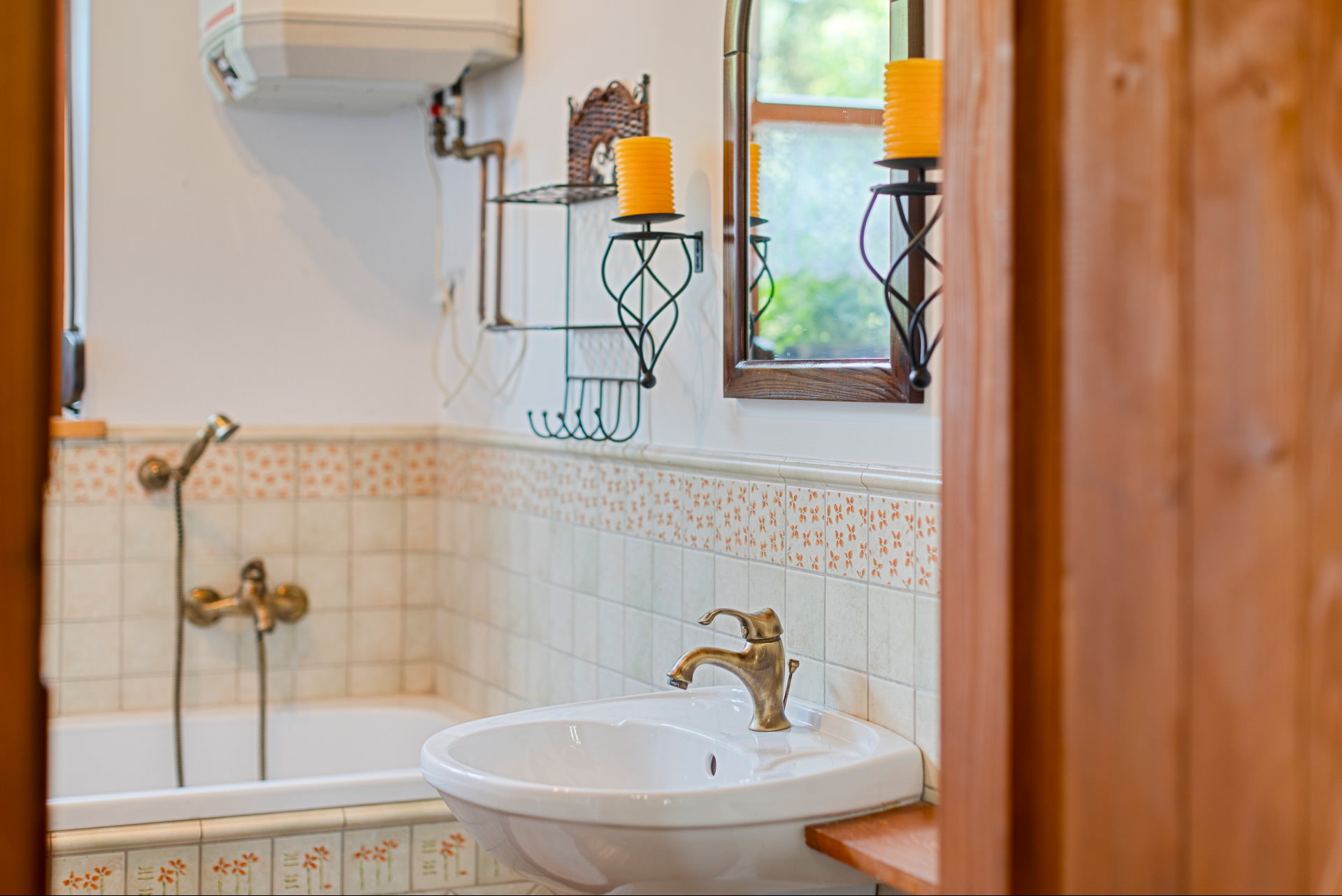 Jasna łazienka z białą umywalką i baterią w stylu retro, widok przez drewniane drzwi. Na ścianie dekoracyjne płytki i lustro z świecami.