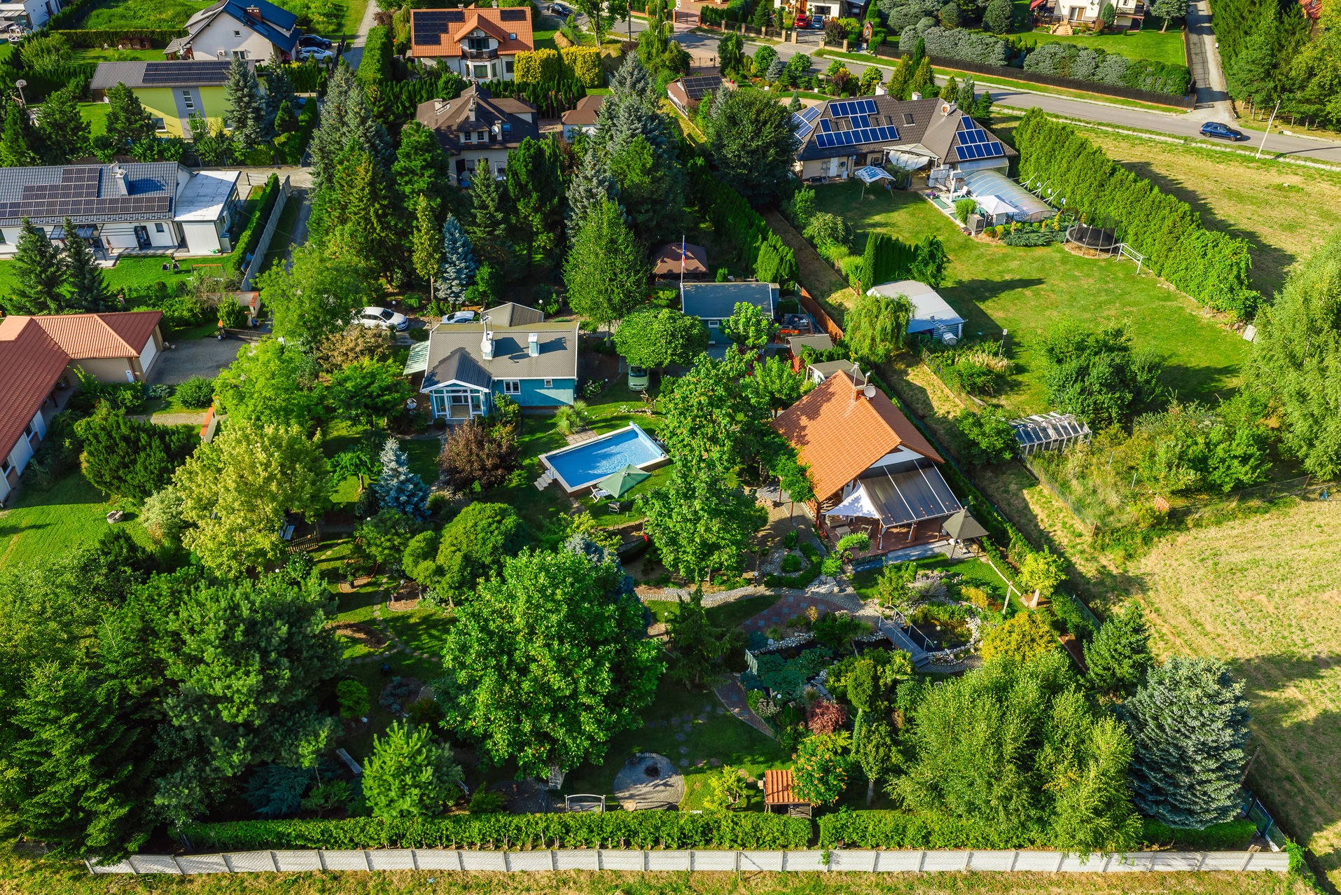 Zdjęcie z lotu ptaka przedstawia osiedle domów jednorodzinnych z basenem, ogrodami i panelami słonecznymi na dachach. Ujęcie w słoneczny dzień.