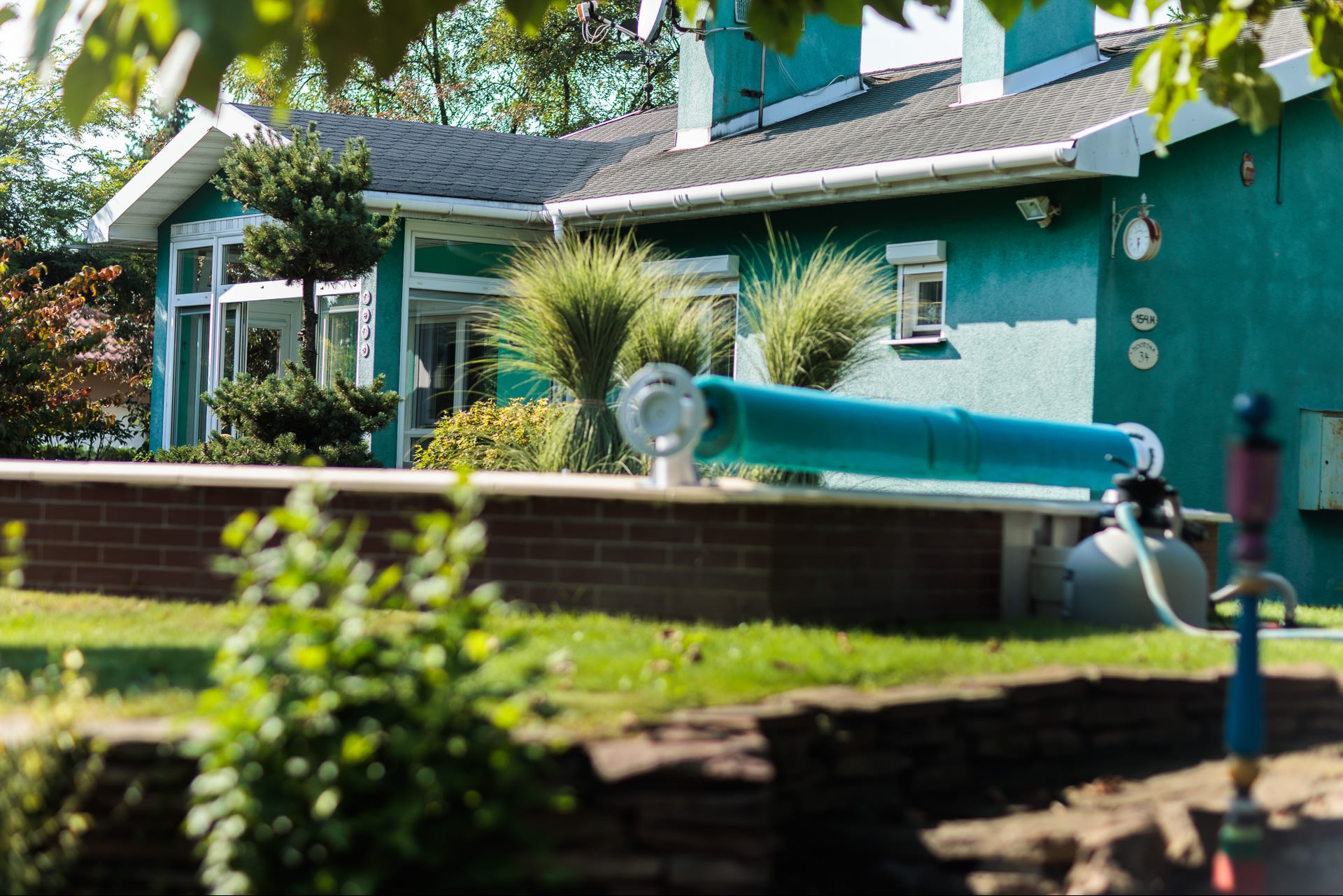 Turkusowy dom z białymi oknami i zielonym ogrodem, widoczny przez roślinność. Na ścianie zegar i tabliczki adresowe. W tle drzewa i niebo.