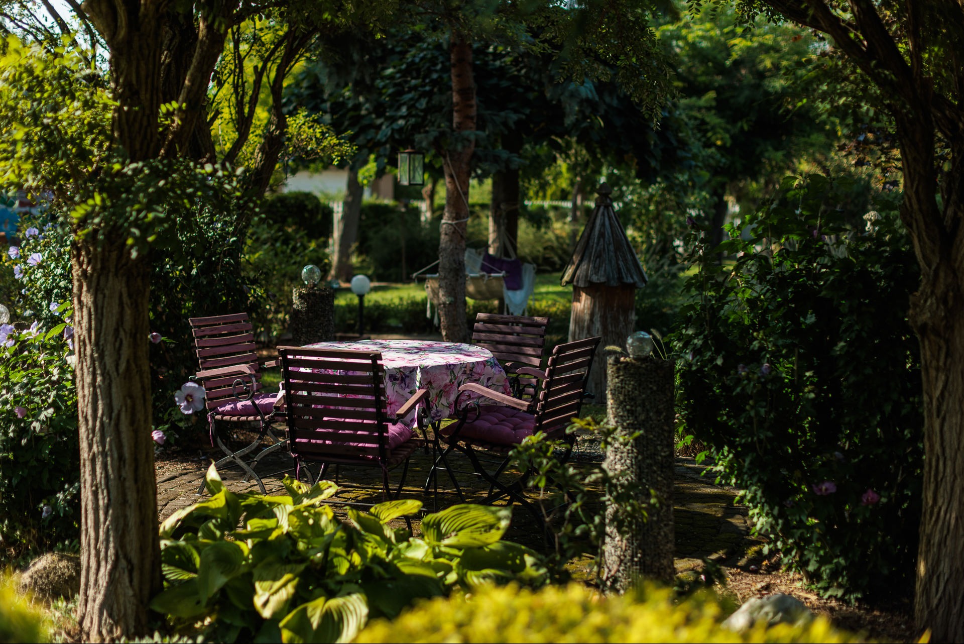 Urokliwy zakątek ogrodu z drewnianym zestawem mebli ogrodowych w odcieniach różu, otoczony bujną zielenią i dekoracyjnymi elementami. Idealne miejsce na relaks.