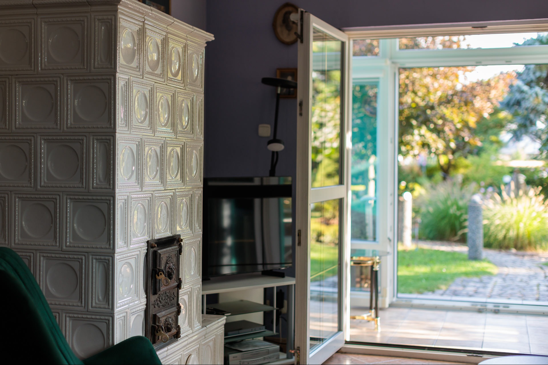 Biały piec kaflowy z ozdobnymi drzwiczkami w salonie z widokiem na ogród przez otwarte drzwi balkonowe. W tle telewizor i meble.