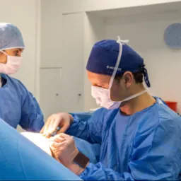 Dwóch chirurgów w niebieskich strojach ochronnych i maskach wykonuje zabieg, używając narzędzi chirurgicznych; widoczny monitor medyczny i zegar na ścianie w tle.