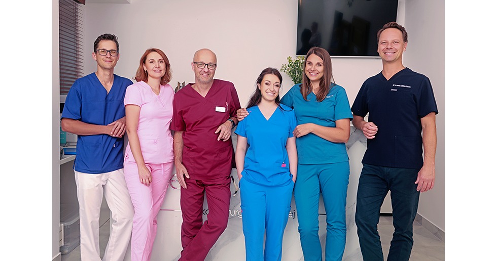 Zespół sześciu uśmiechniętych lekarzy w kolorowych uniformach pozuje w jasnym wnętrzu kliniki z telewizorem i kwiatami w tle.