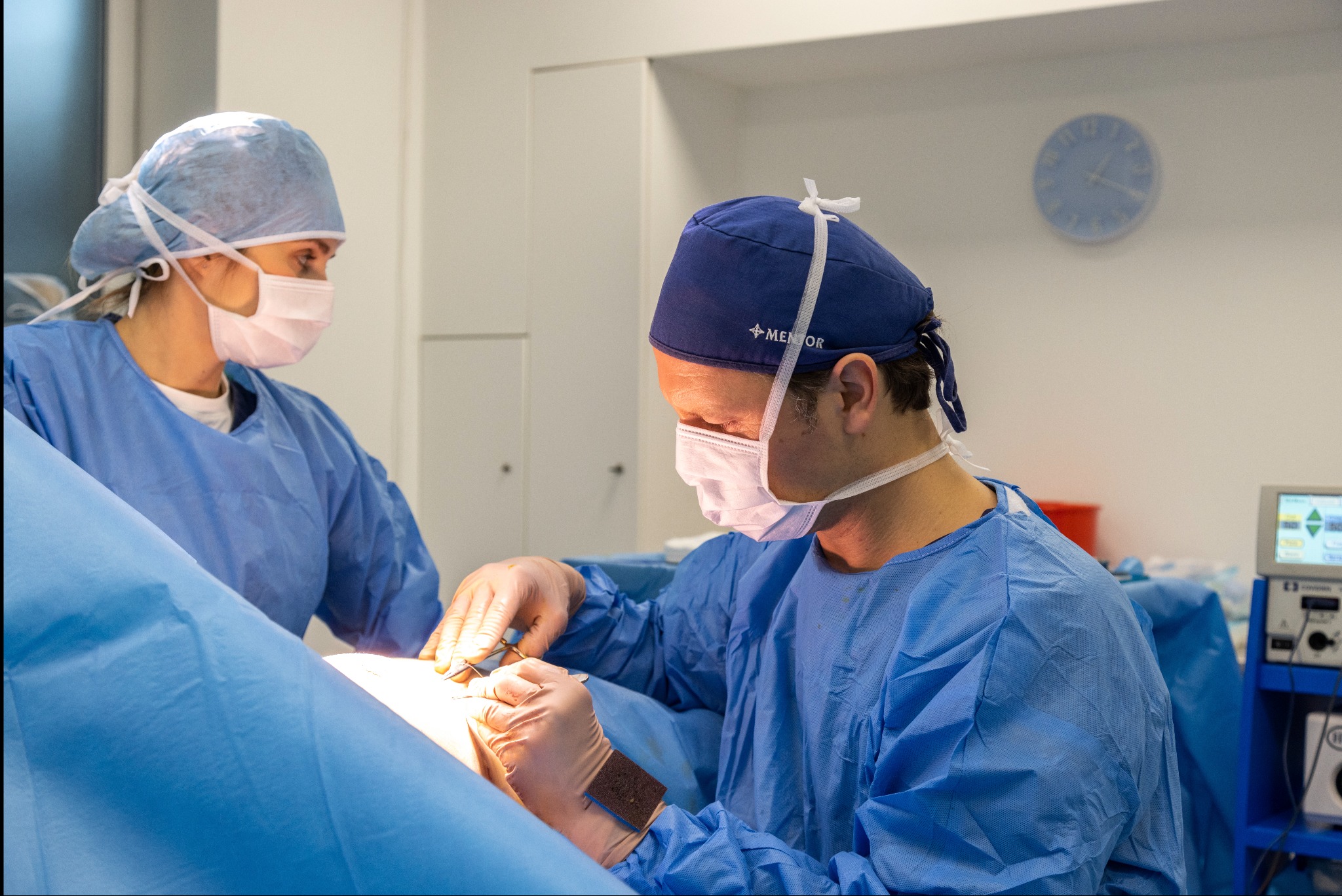 Dwóch chirurgów w niebieskich strojach ochronnych i maskach wykonuje zabieg, używając narzędzi chirurgicznych; widoczny monitor medyczny i zegar na ścianie w tle.