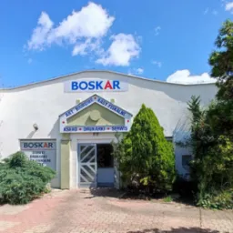 Fasada budynku firmy BOSKAR z szyldami informującymi o sprzedaży artykułów biurowych, kas fiskalnych, kserokopiarek, drukarek i serwisie. Budynek otoczony zielenią, na pierwszym planie kostka brukowa.