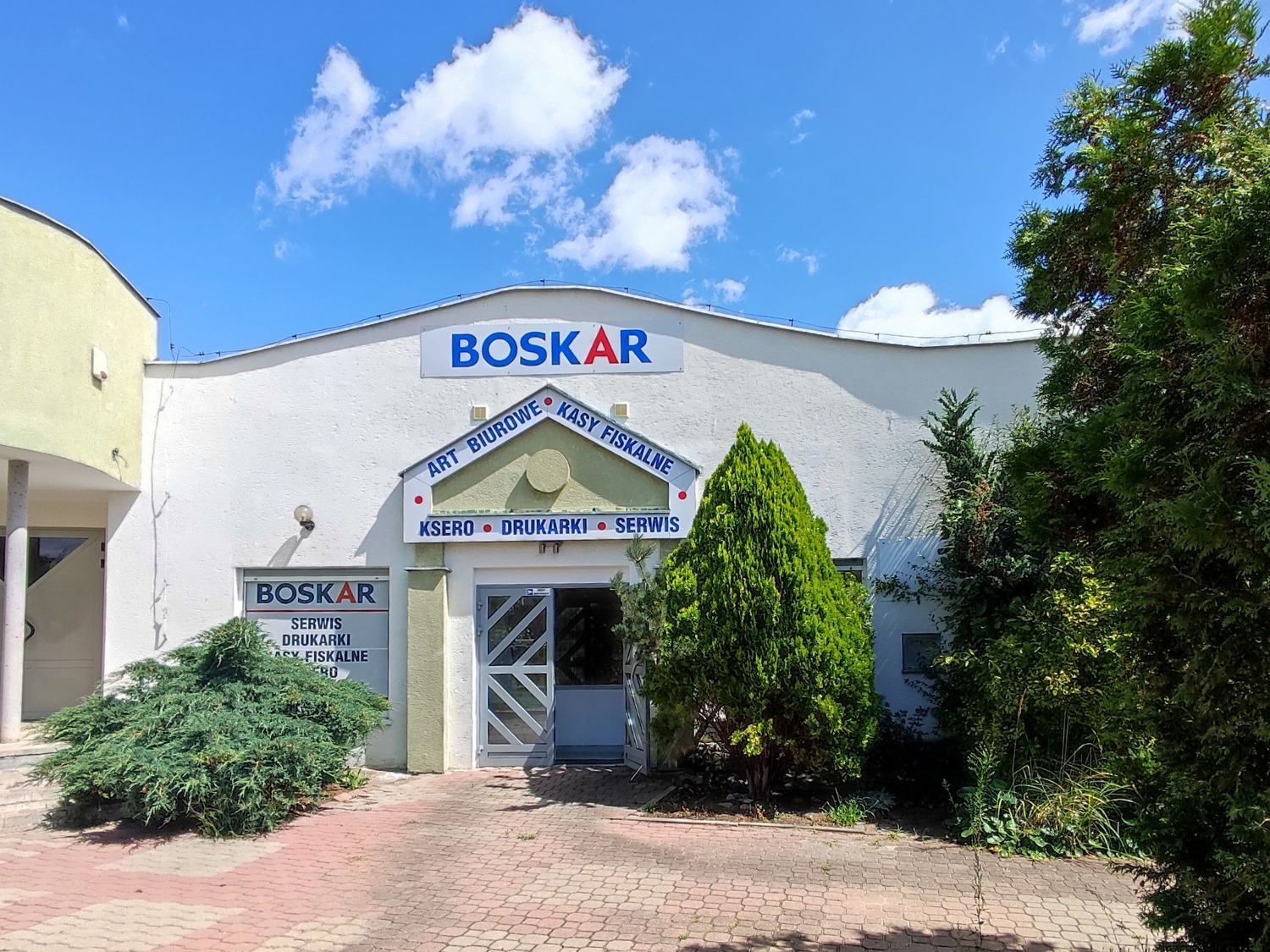 Fasada budynku firmy BOSKAR z szyldami informującymi o sprzedaży artykułów biurowych, kas fiskalnych, kserokopiarek, drukarek i serwisie. Budynek otoczony zielenią, na pierwszym planie kostka brukowa.