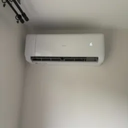 Nowoczesny, biały klimatyzator ścienny marki ANDE z wyświetlaną temperaturą 16 stopni, zamontowany w rogu jasnego pomieszczenia z zasłonami w tle.