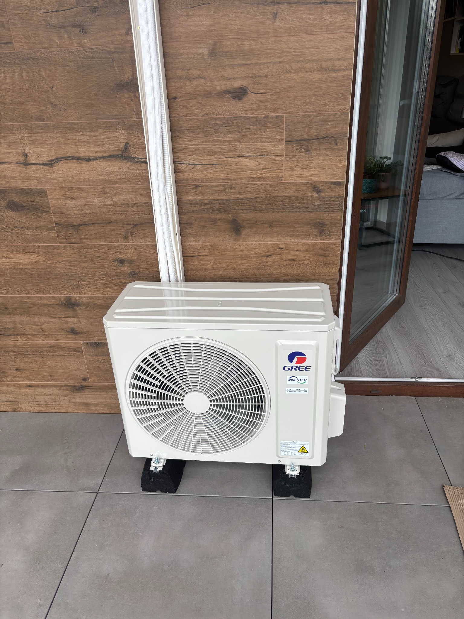 Biały klimatyzator Gree Inverter na zewnątrz, na szarych płytkach, z brązową ścianą i otwartym oknem balkonowym w tle. Widoczne czarne podkładki pod urządzeniem.