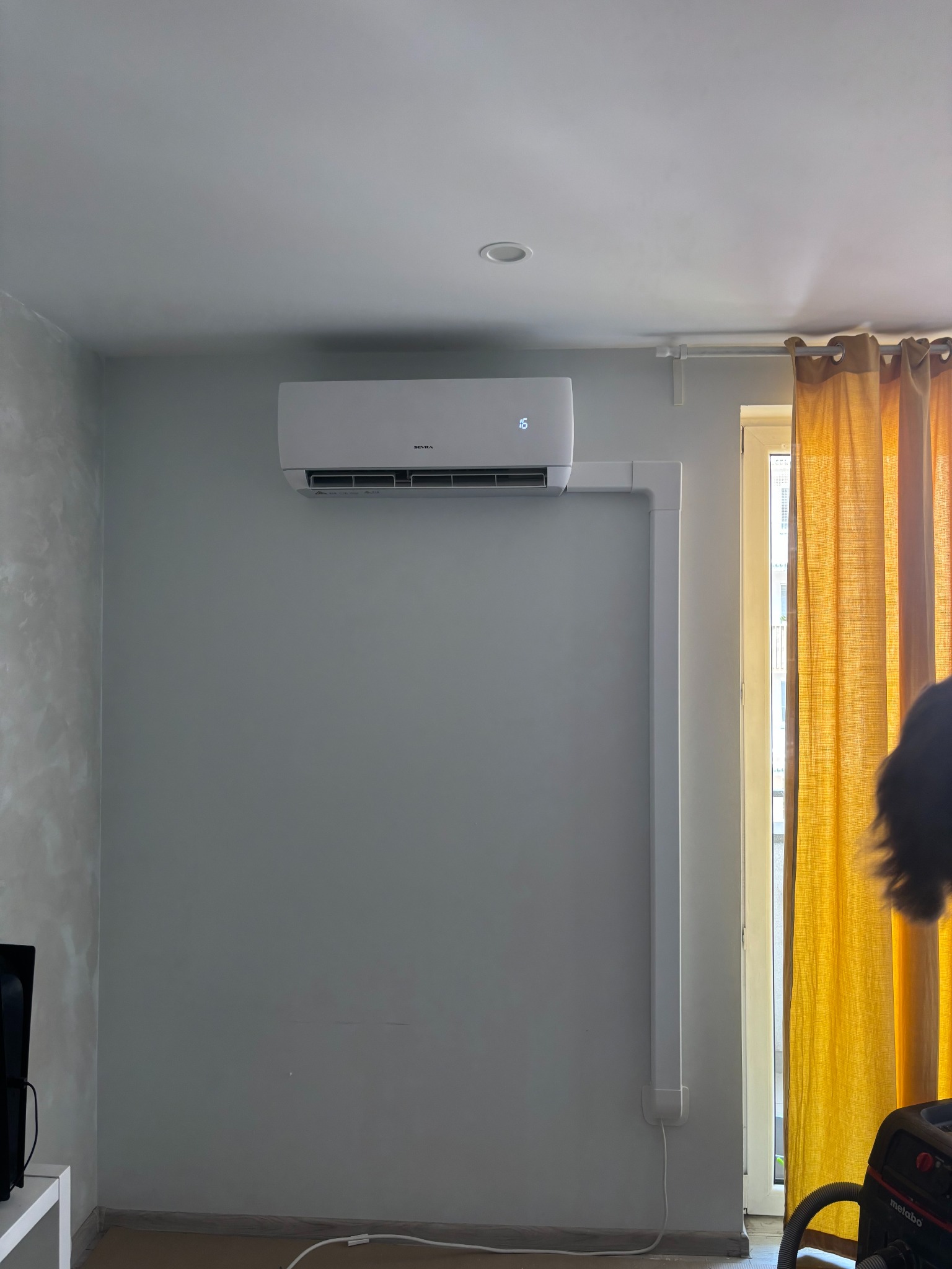 Nowoczesny klimatyzator ścienny z białymi rurami, zamontowany w jasnym pomieszczeniu z żółtymi zasłonami. Widoczny fragment odkurzacza.