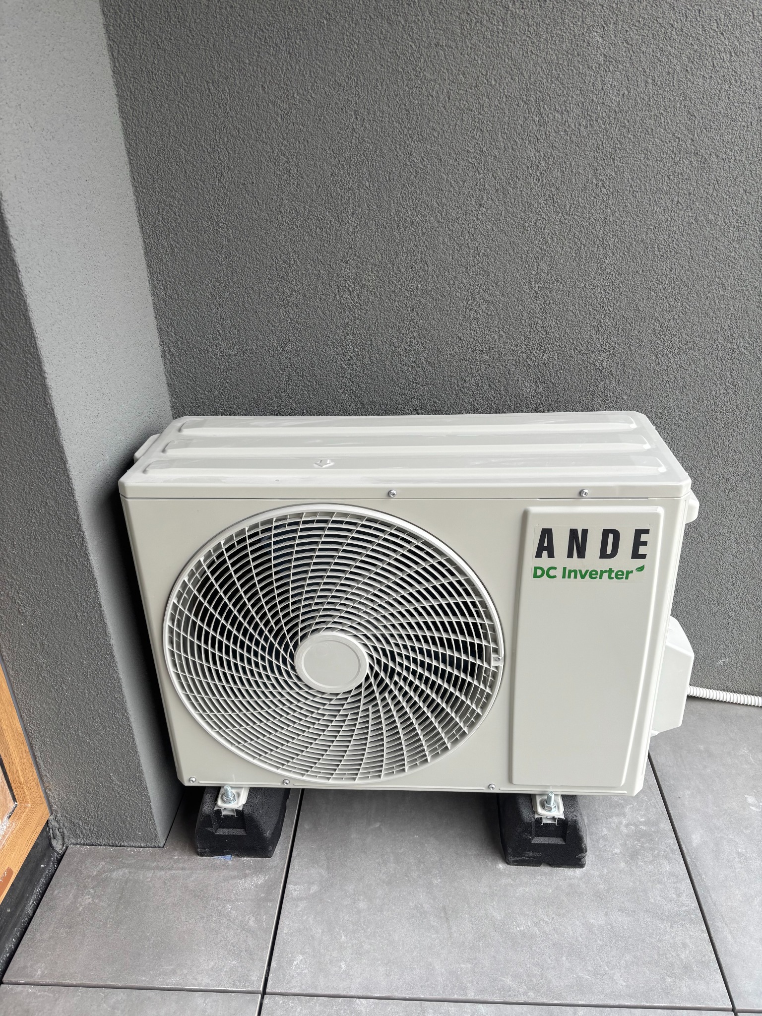 Zamontowana jednostka zewnętrzna klimatyzacji ANDE DC Inverter, umieszczona na czarnych podkładkach na szarym, kafelkowym balkonie, przy szarej ścianie.