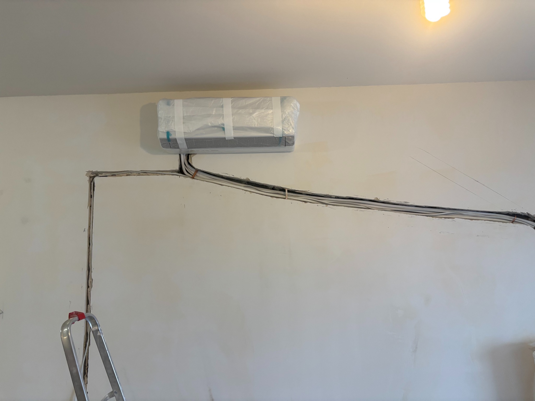 Montaż klimatyzacji: jednostka wewnętrzna zabezpieczona folią na ścianie, widoczne kable w bruździe, drabina. Etap instalacji przed wykończeniem ściany.