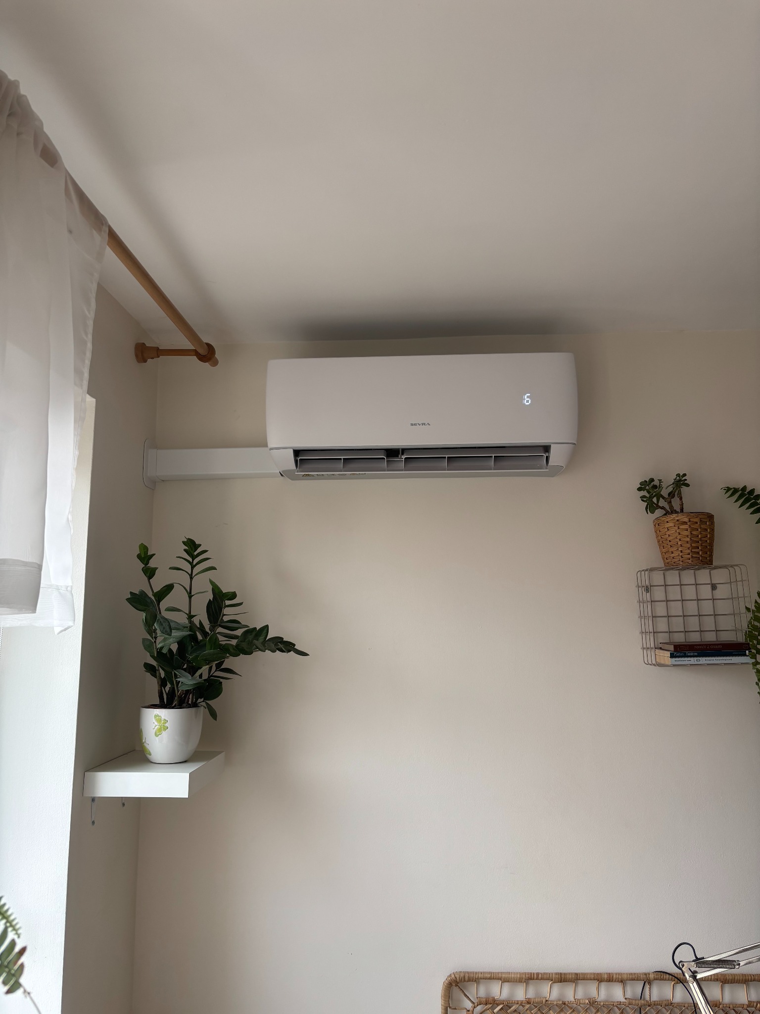 Biały klimatyzator na ścianie w jasnym pokoju, obok rośliny doniczkowe na półkach. Widoczny minimalistyczny design urządzenia i otoczenia.
