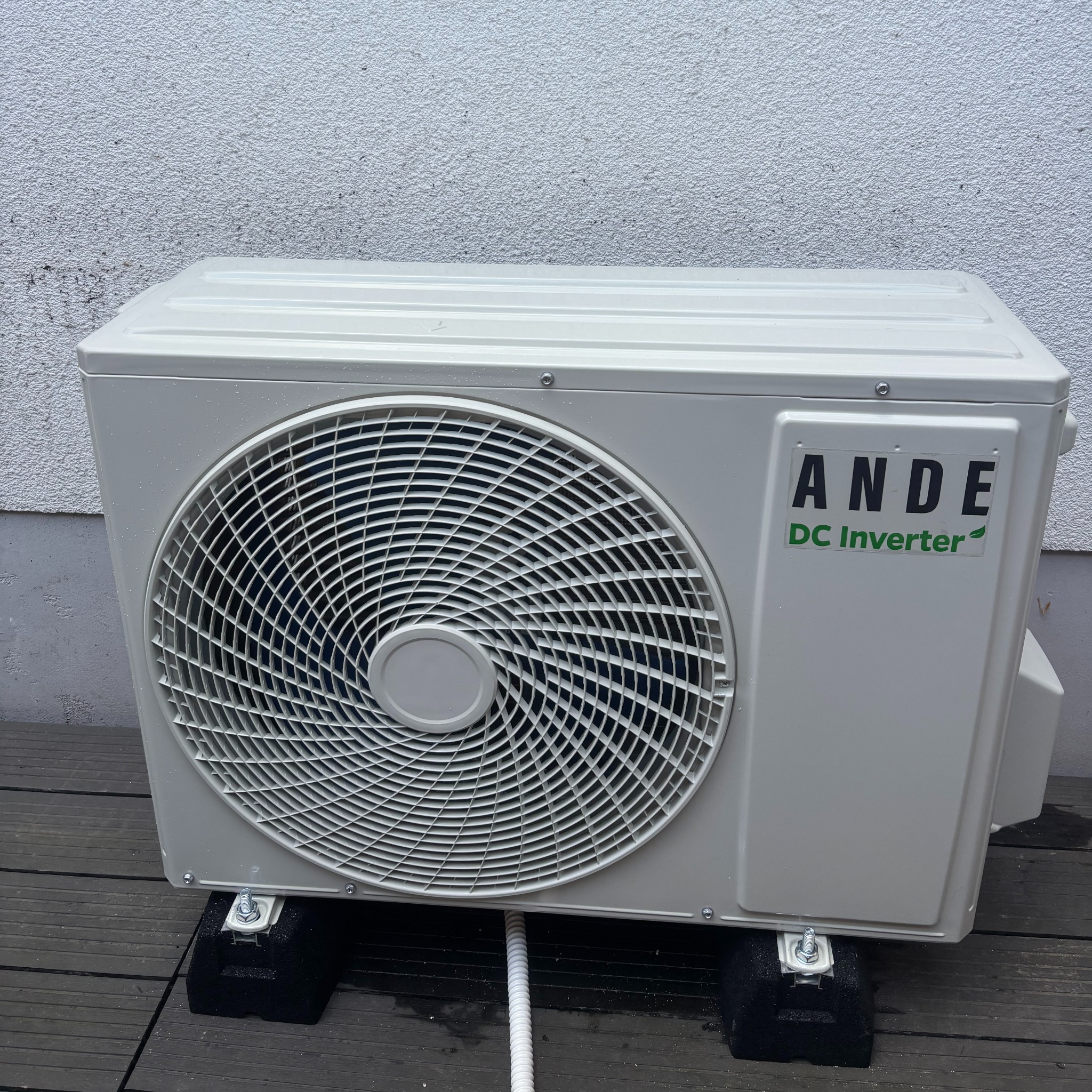 Zewnętrzna jednostka klimatyzacji ANDE DC Inverter, zamontowana na czarnych podkładkach, z widoczną białą rurą odprowadzającą skropliny na drewnianym tarasie.