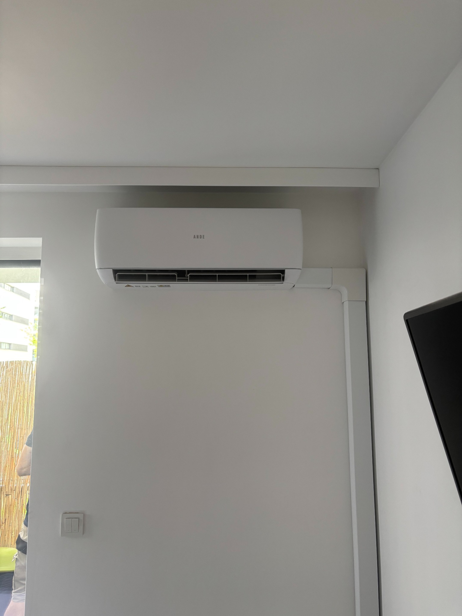 Biały klimatyzator ANDE zamontowany na ścianie w jasnym pomieszczeniu. Widoczne białe maskownice instalacji elektrycznej. Fragment okna i telewizora w kadrze.