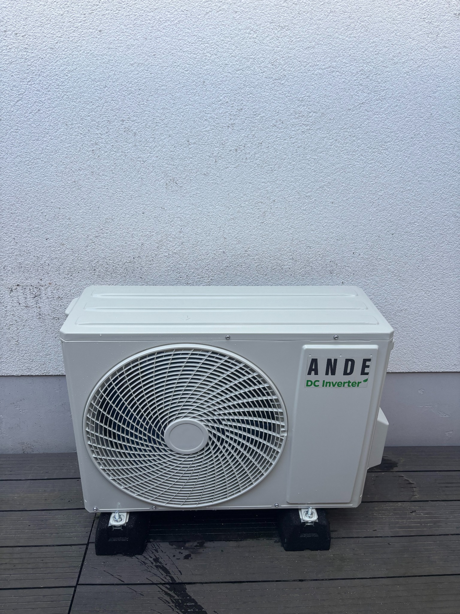 Biała jednostka zewnętrzna klimatyzacji ANDE DC Inverter, ustawiona na czarnych podkładkach na drewnianym tarasie, na tle białej ściany. Widoczne detale montażowe.