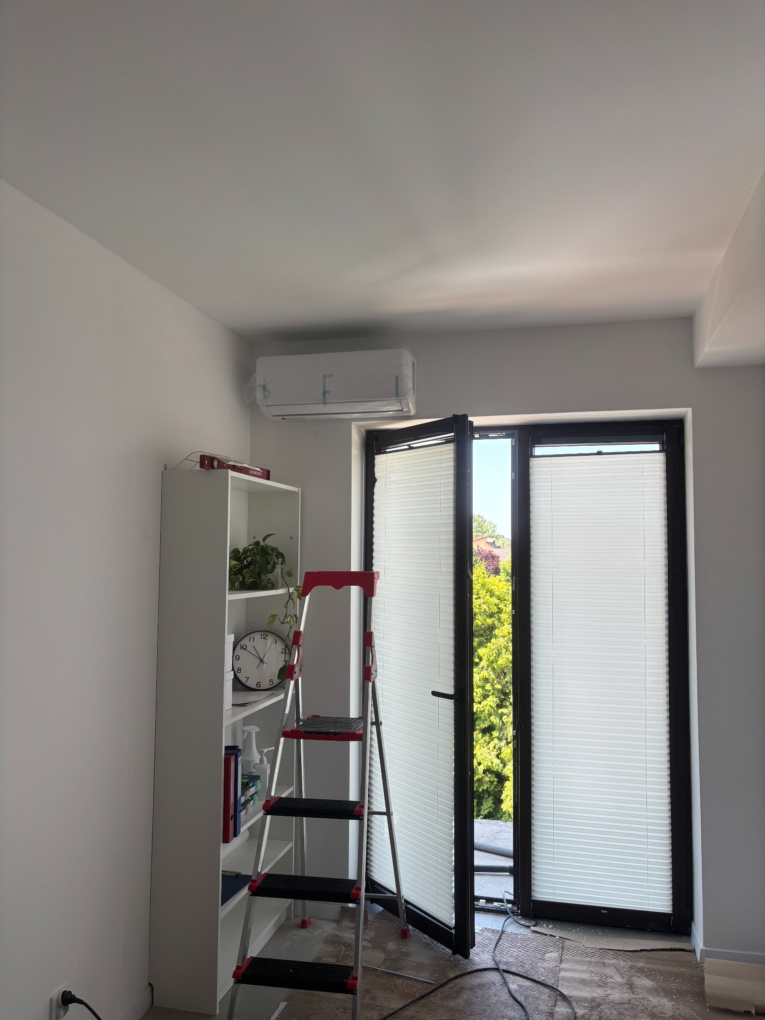 Biały klimatyzator na ścianie nad oknem z żaluzjami plisowanymi. Obok regał i drabina, widoczne przewody. Montaż klimatyzacji w domu.