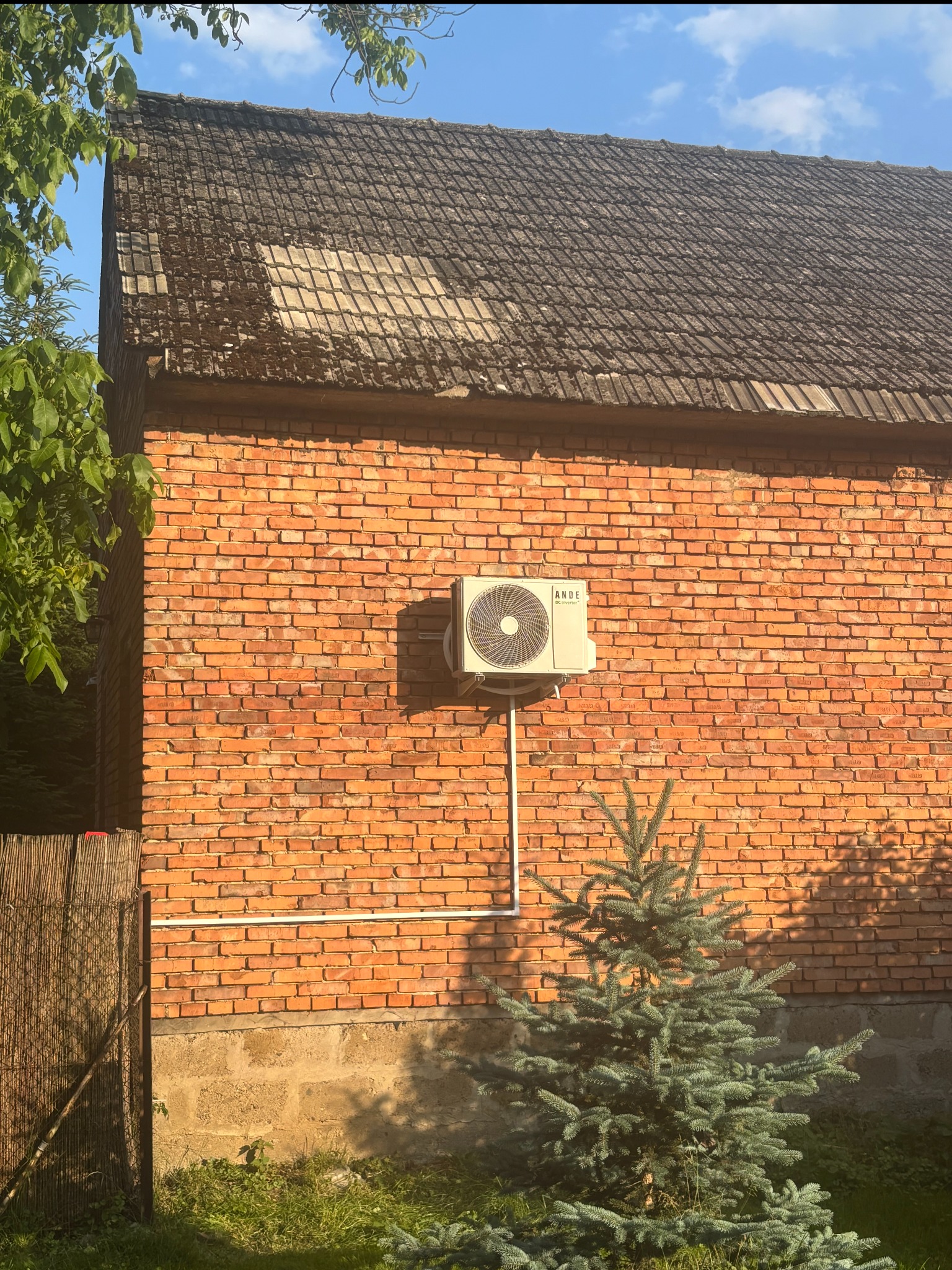 Zamontowana klimatyzacja na ceglanej ścianie budynku z widocznym przewodem. W tle dach pokryty starą dachówką i fragment zieleni.