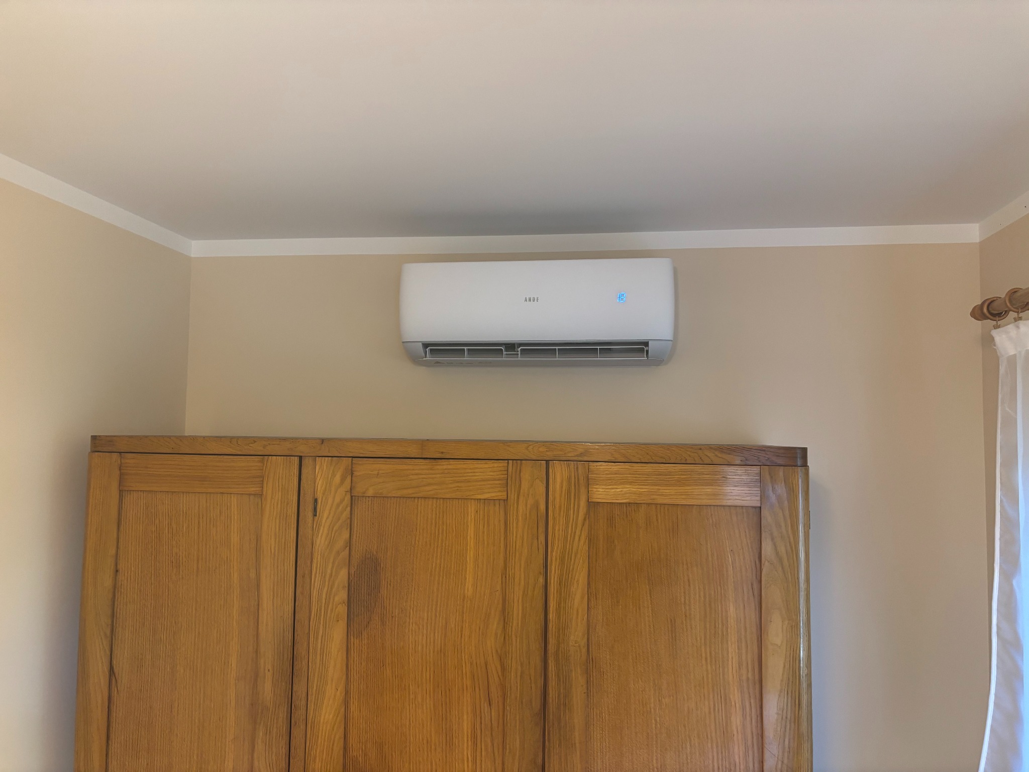 Biały klimatyzator ścienny marki ANDE zamontowany nad drewnianą szafą w pokoju o beżowych ścianach i białym suficie. Widoczny fragment zasłony.