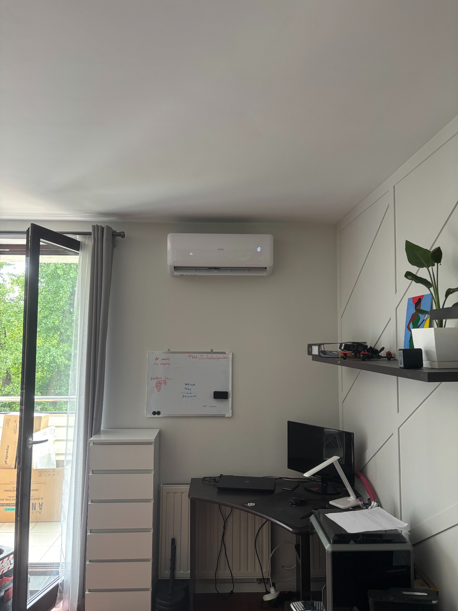 Klimatyzator ścienny zamontowany w pokoju z biurkiem, półką i otwartym balkonem. Nowoczesny design, jasne wnętrze, widok na zieleń za oknem.