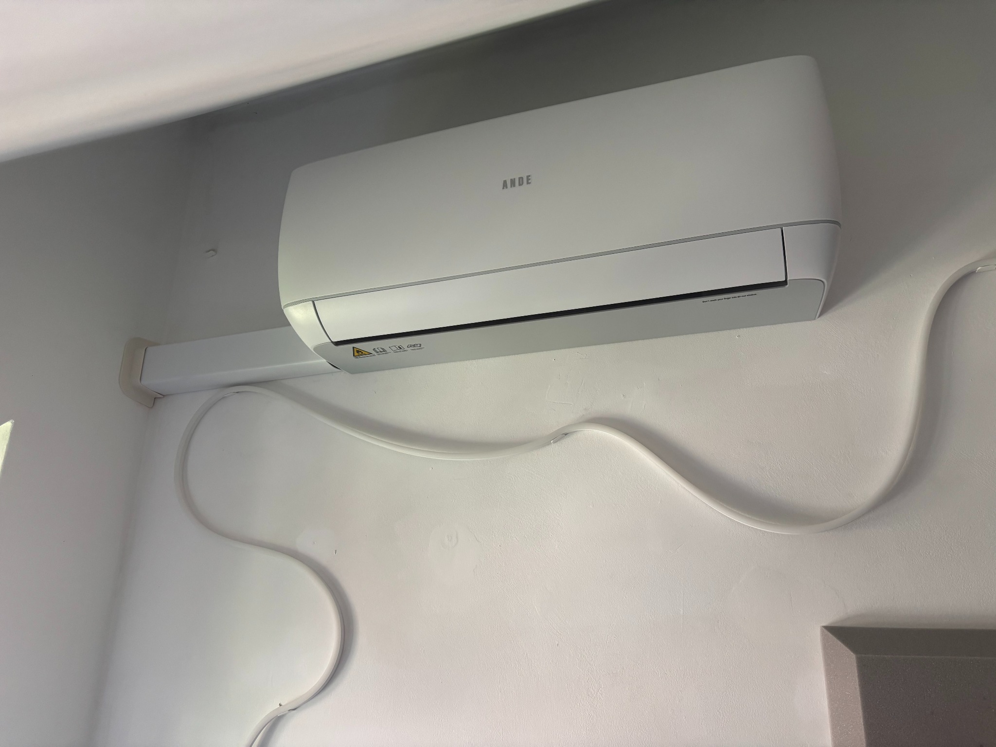 Biała klimatyzacja ANDE zamontowana na białej ścianie, widoczne przewody. Minimalistyczny design, czyste linie, oświetlenie dzienne.