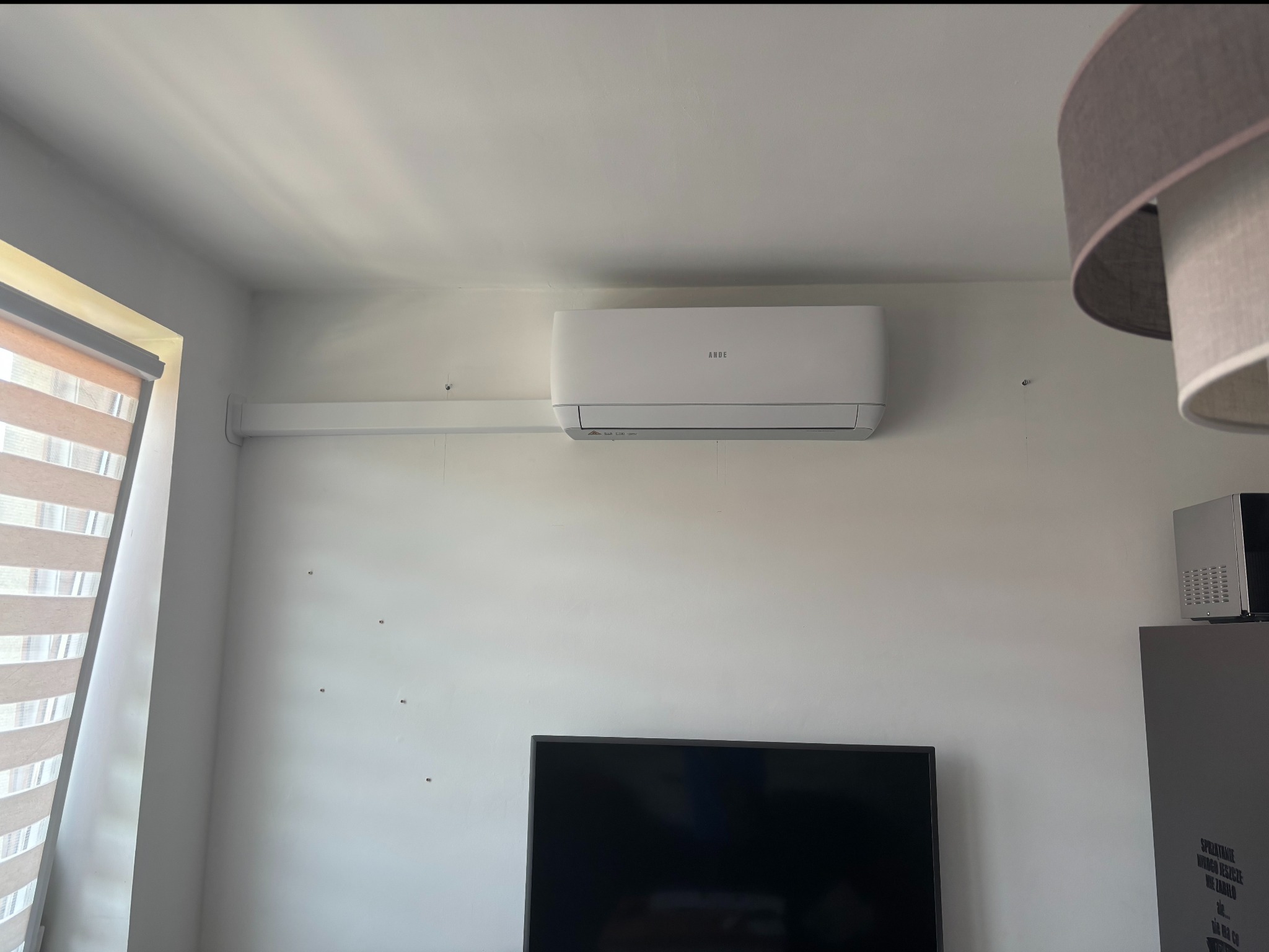 Biały klimatyzator ścienny marki ANDE zamontowany w jasnym pokoju. Widoczne mocowania na ścianie, okno z żaluzjami i fragment lampy.