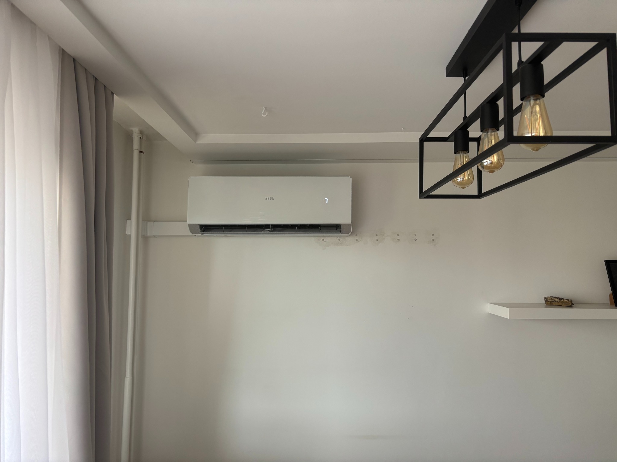 Biały klimatyzator na ścianie w jasnym wnętrzu, widoczny fragment zasłony i nowoczesna lampa sufitowa z trzema żarówkami, schludne wykończenie.