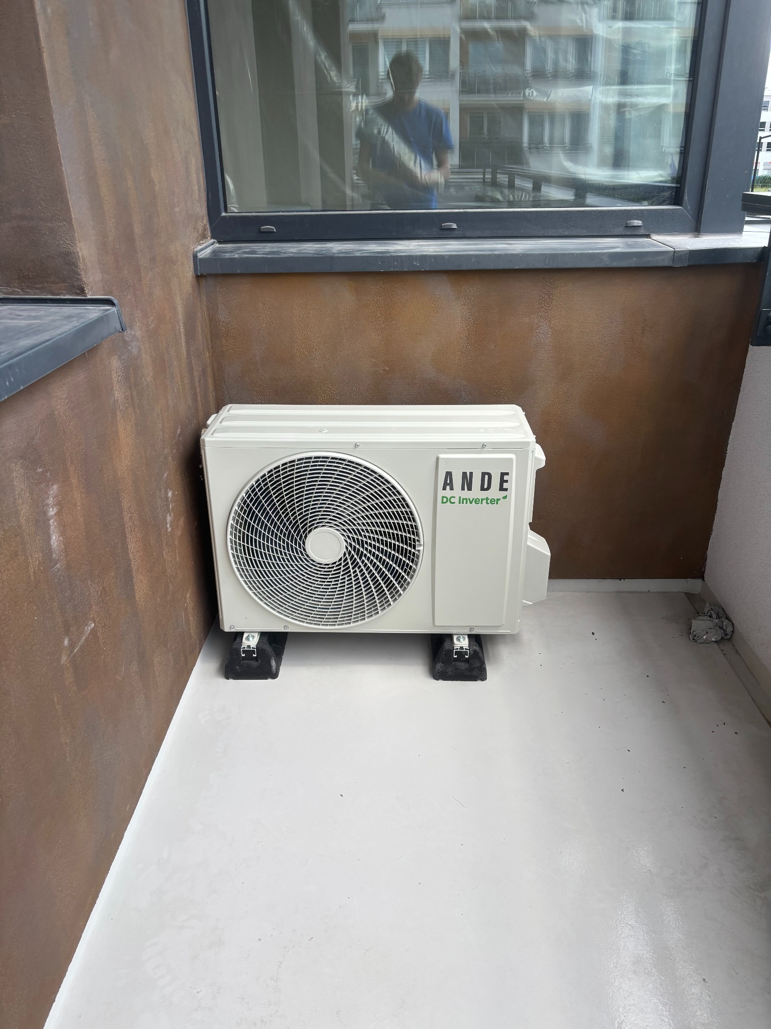 Kompaktowa klimatyzacja ANDE DC Inverter, umieszczona na balkonie z brązową ścianą i dużym oknem, zapewniająca komfortowe warunki w mieszkaniu.