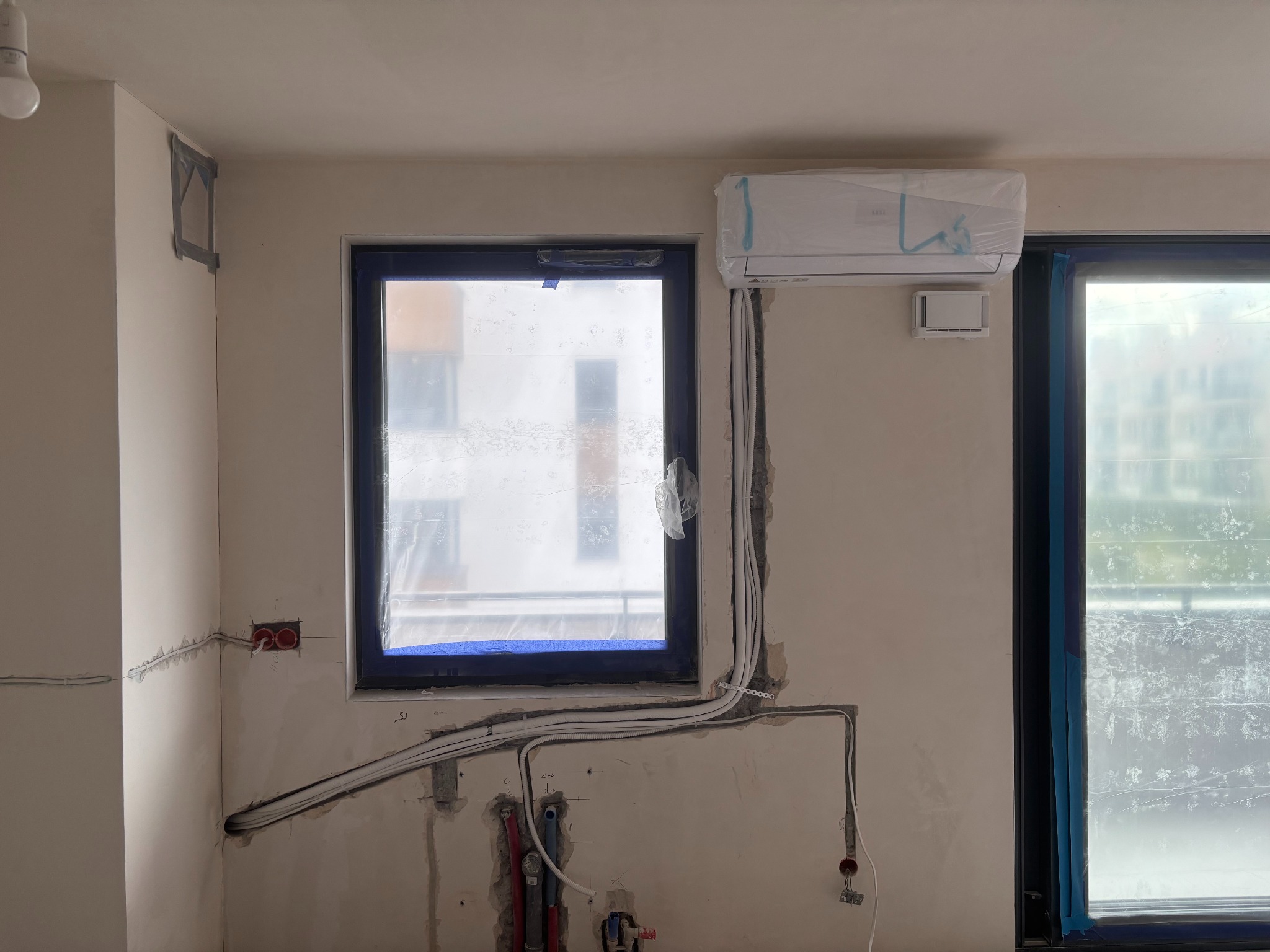 Montaż klimatyzacji w surowym wnętrzu: jednostka wewnętrzna zabezpieczona folią, widoczne przewody i instalacje przed wykończeniem ścian. Okno z niebieską taśmą.