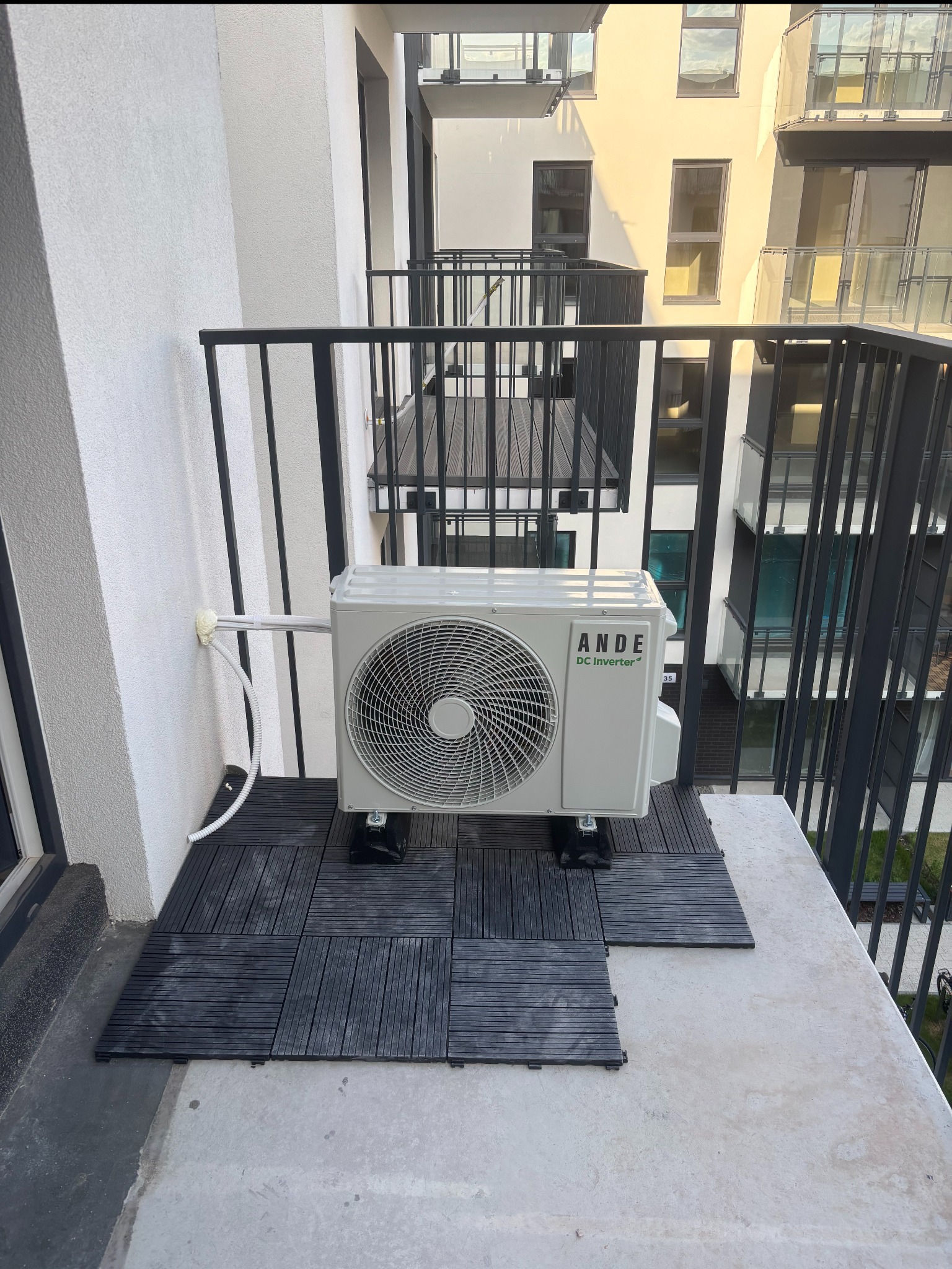 Biała jednostka zewnętrzna klimatyzacji ANDE DC Inverter na balkonie nowoczesnego apartamentowca, widok z góry, czarne płytki pod urządzeniem.