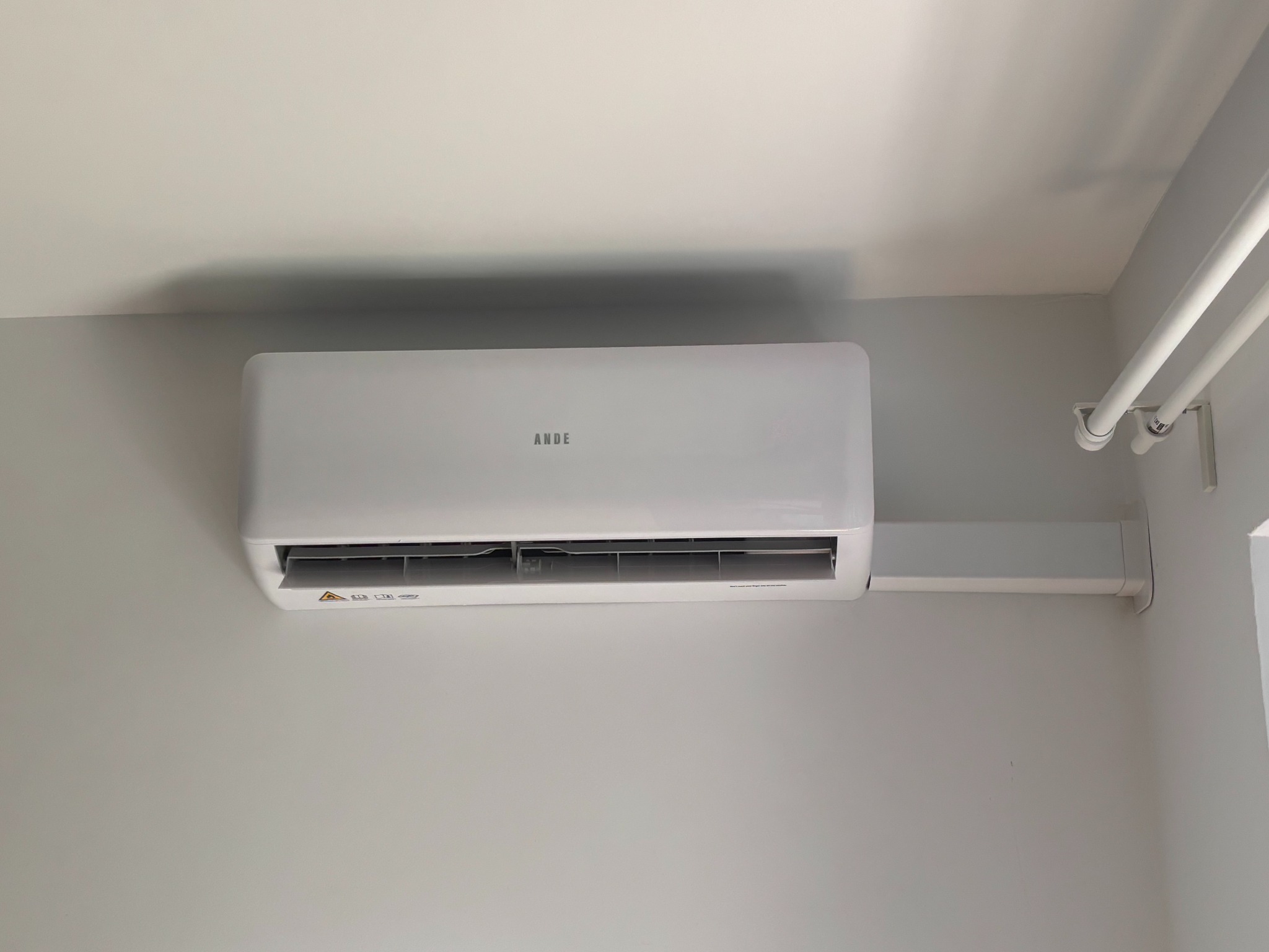 Biała klimatyzacja ANDE zamontowana na szarej ścianie w pomieszczeniu. Widoczne rury instalacyjne przy suficie. Ujęcie z dołu, lekko z prawej strony.