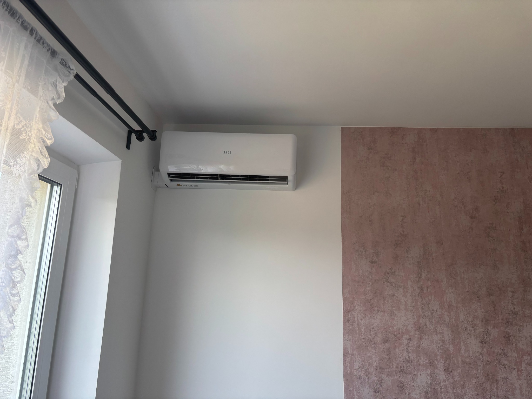 Biały klimatyzator ANDE zamontowany na białej ścianie w pokoju z oknem z firanką i fragmentem ściany z różową tapetą. Widok z dołu.
