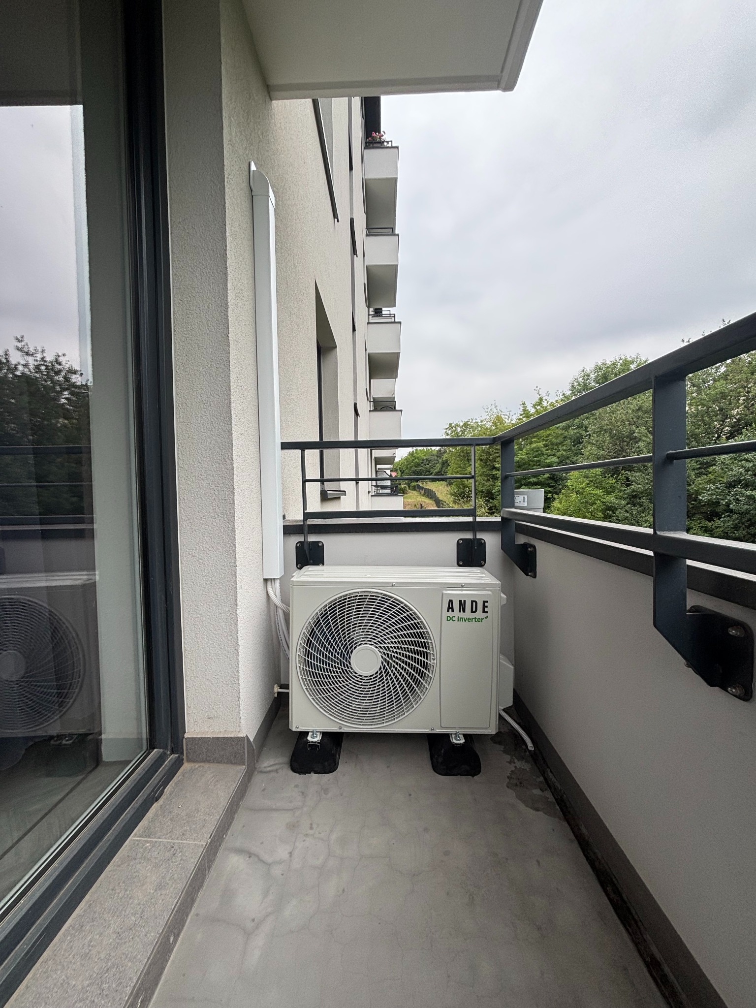 Biała jednostka zewnętrzna klimatyzacji ANDE DC Inverter na balkonie w Krakowie. Widok na szarą balustradę i zieleń drzew w tle. Montaż przy ścianie budynku.