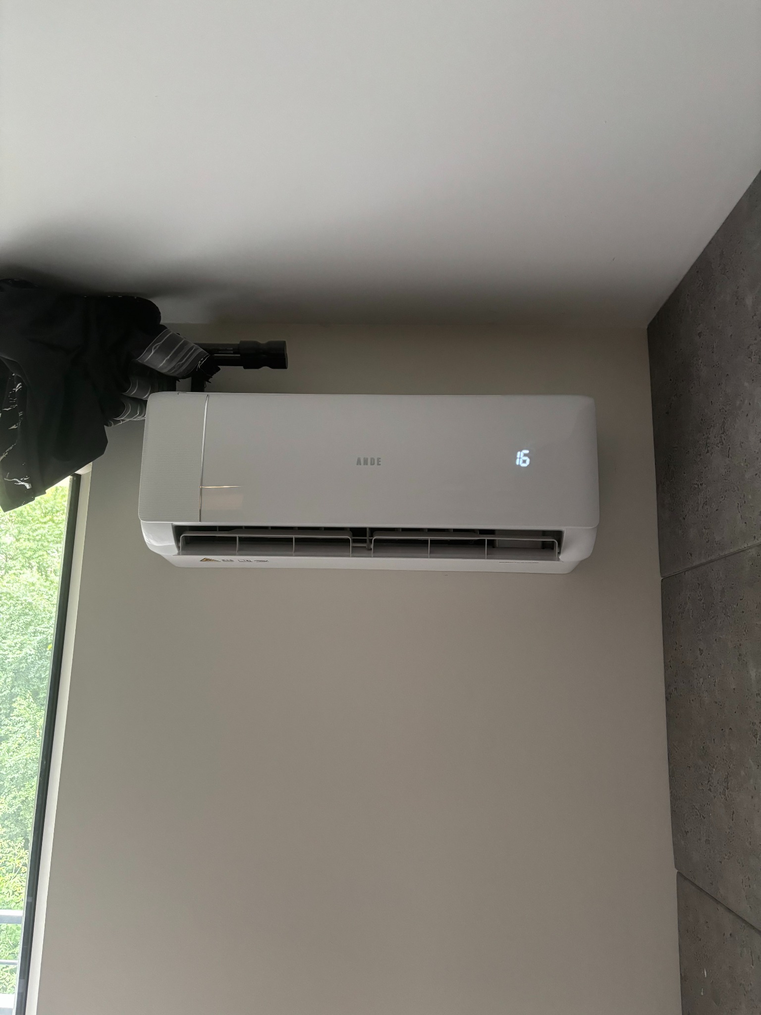Biały klimatyzator ANDE z wyświetlaną temperaturą 16 stopni Celsjusza, zamontowany na ścianie w pomieszczeniu z widokiem na zieleń za oknem.