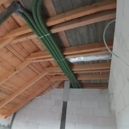 Grzegorz Dudzik - Zielone rury wentylacyjne przymocowane do drewnianej konstrukcji dachu na poddaszu, z widoczną izolacją termiczną i fragmentem ściany z bloczków betonowych.